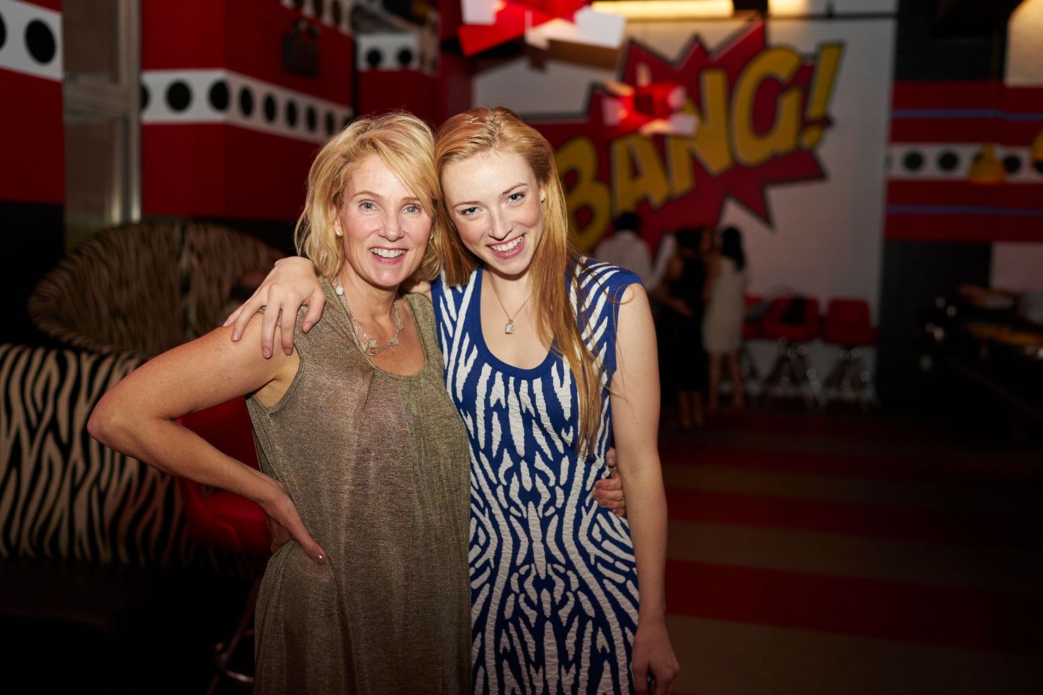 Two friends pose together at a party venue with red and white decor visible in the background.