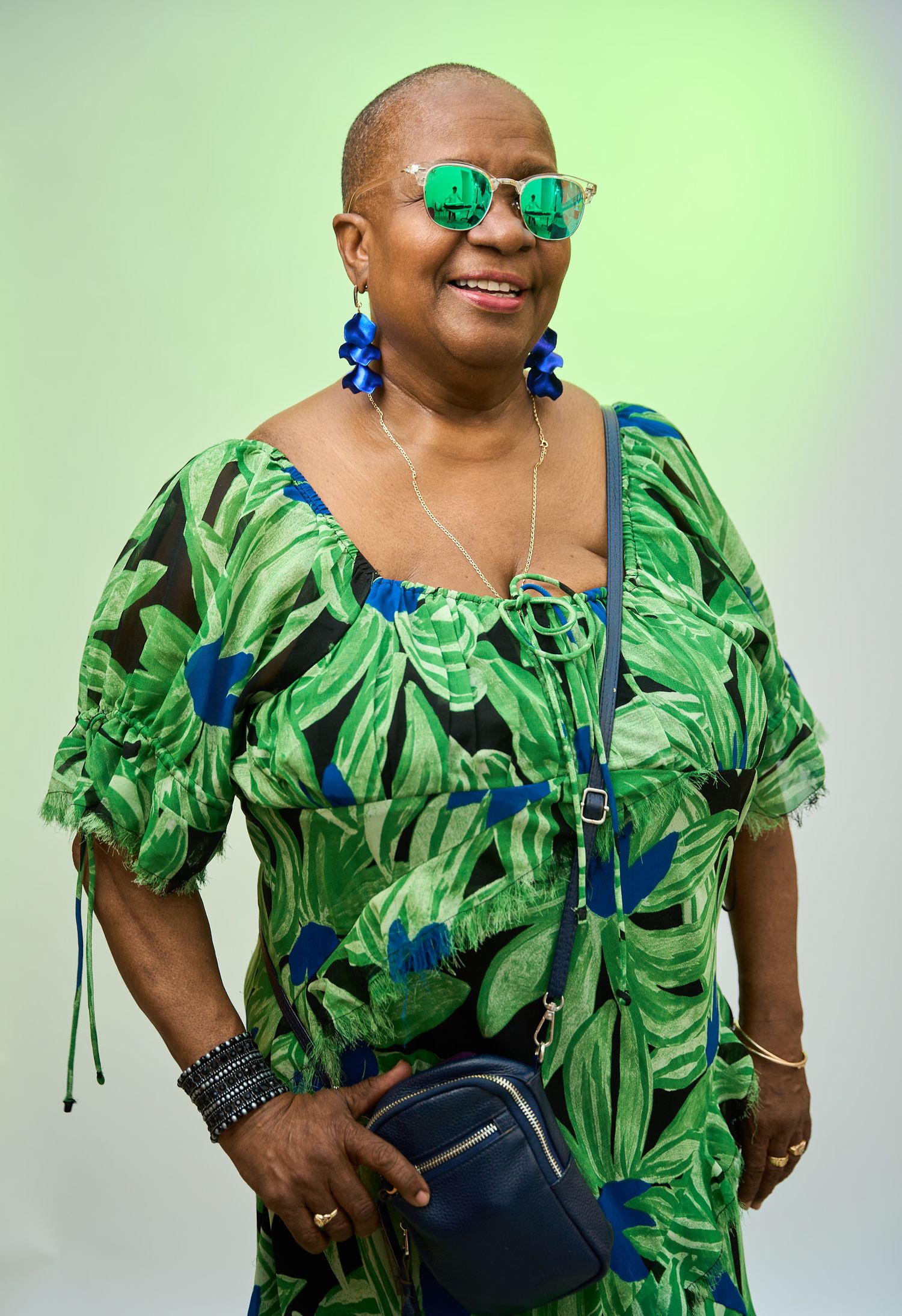 Smiling older woman in green floral dress, blue earrings, sunglasses, holding navy purse against light green background.