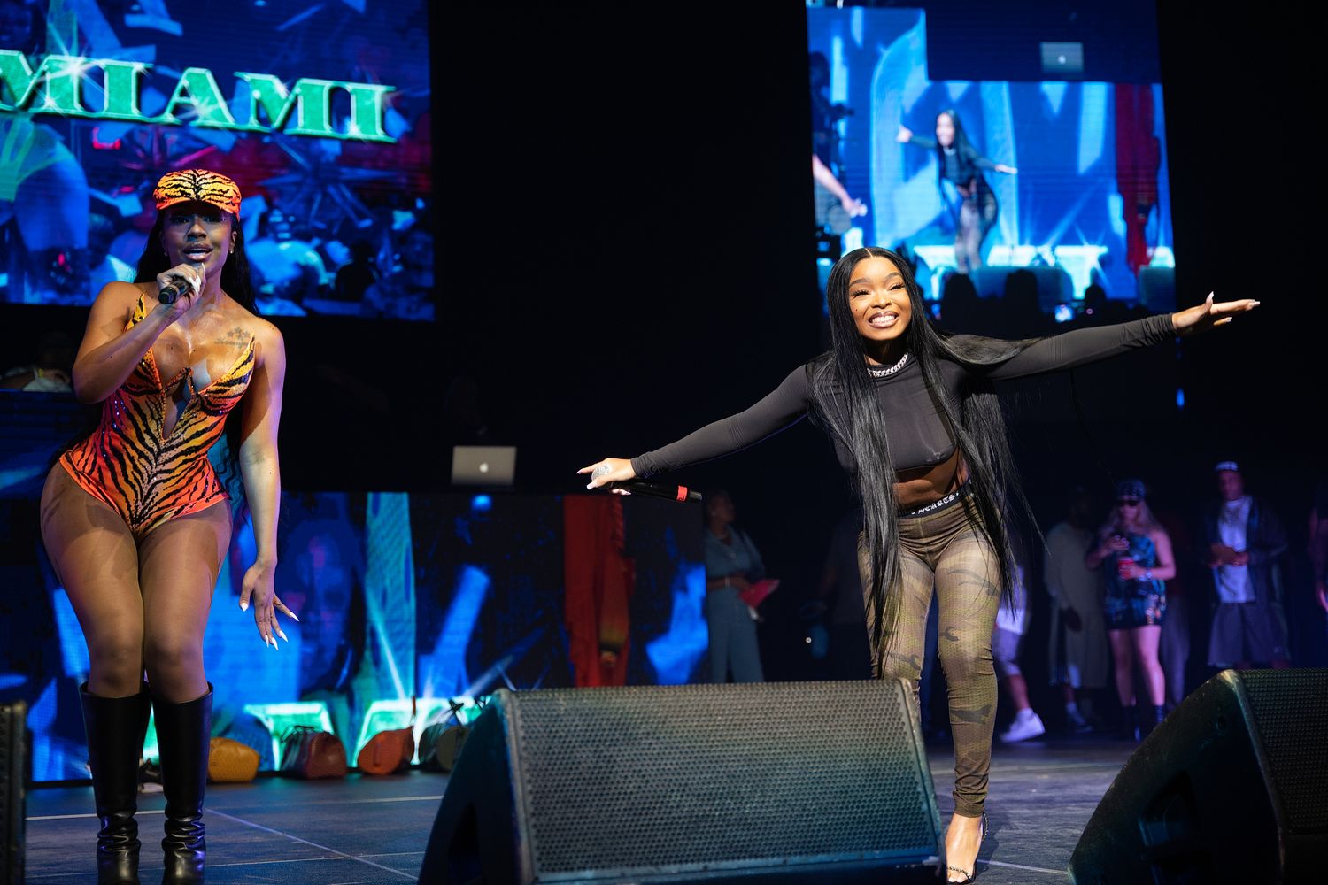 Two female performers on stage in Miami, one in colorful bodysuit with mic, other in black outfit with arms outstretched.