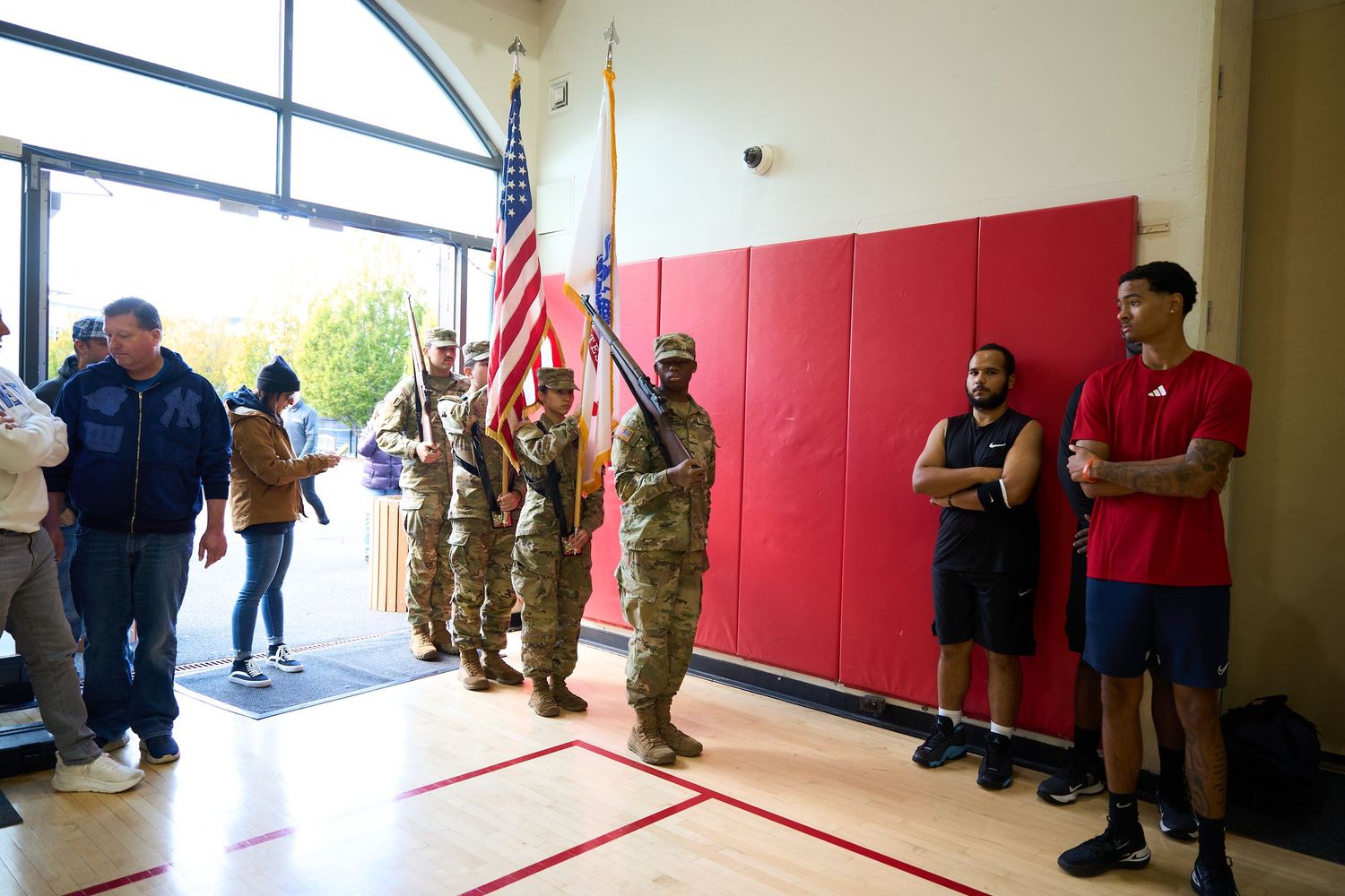 Military personnel and civilians gather along red walls in a gymnasium hallway.