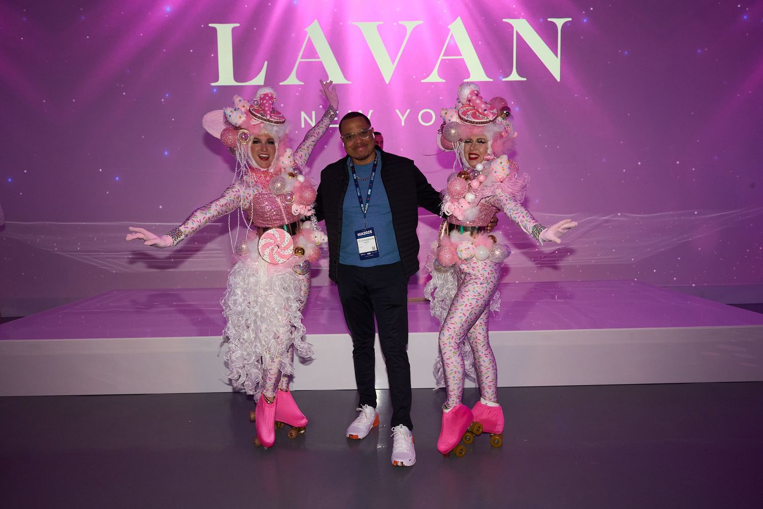 Models walk the runway in pink and white outfits during a fashion show with purple lighting and stage effects.