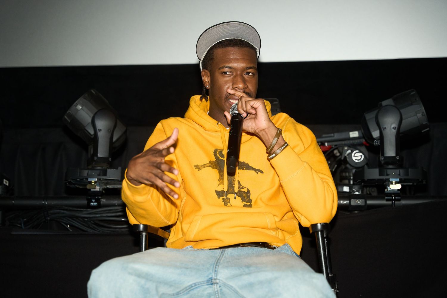 Young man in a yellow hoodie and gray cap sits on stage speaking into a microphone at an indoor event.