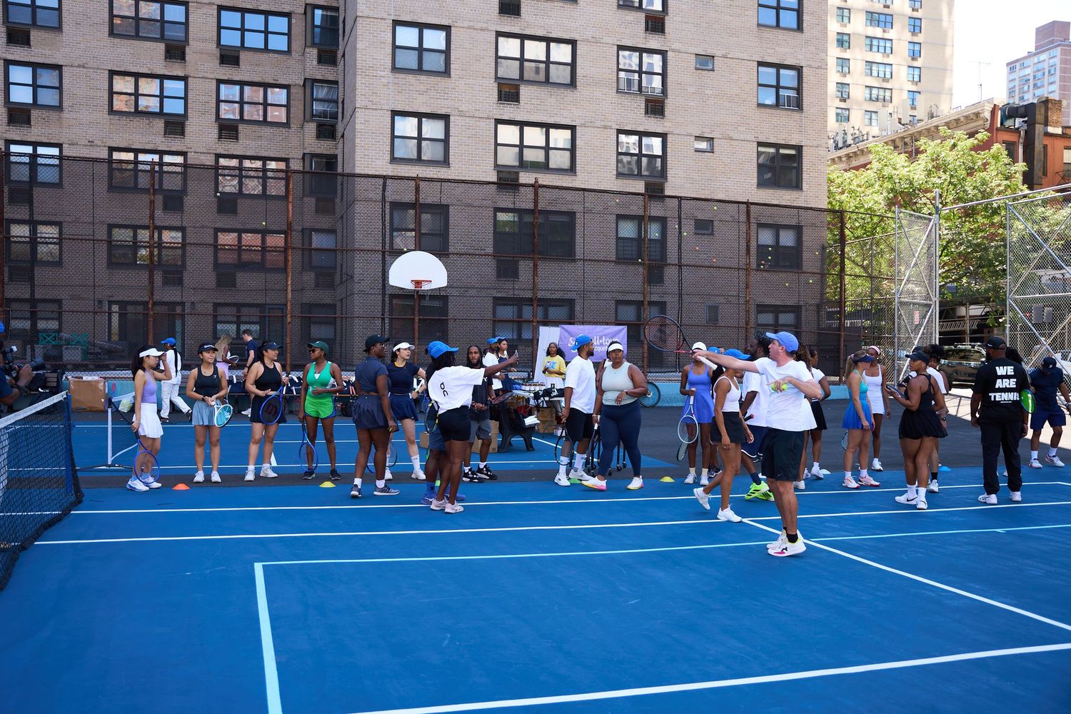 Players compete in a pickleball match on a bright blue outdoor court surrounded by urban buildings.