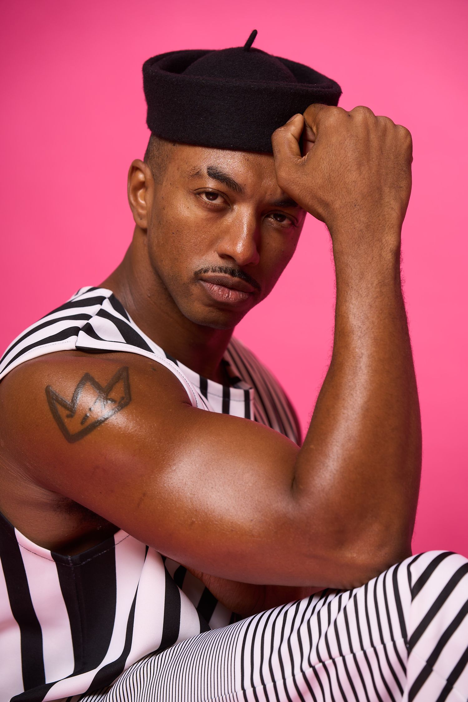 A person wearing a black beret and striped outfit poses against a pink background with one hand near their forehead.