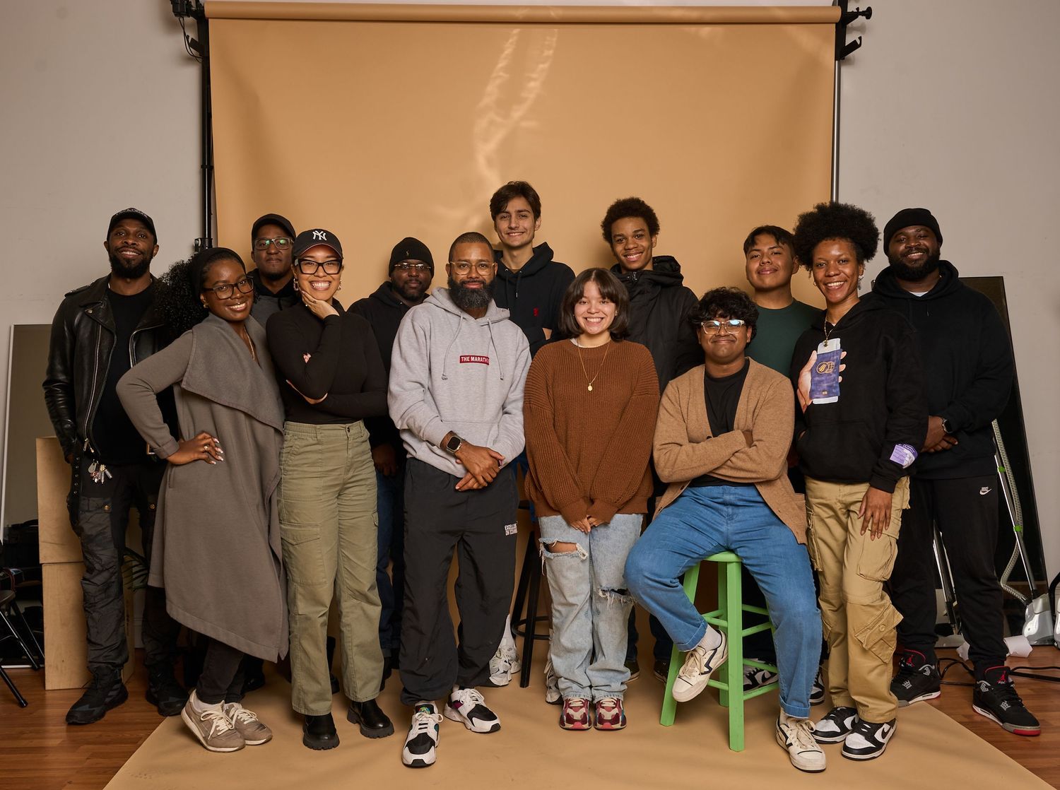 Group of people seated casually against a beige wall wearing sneakers and casual clothing in an indoor setting.