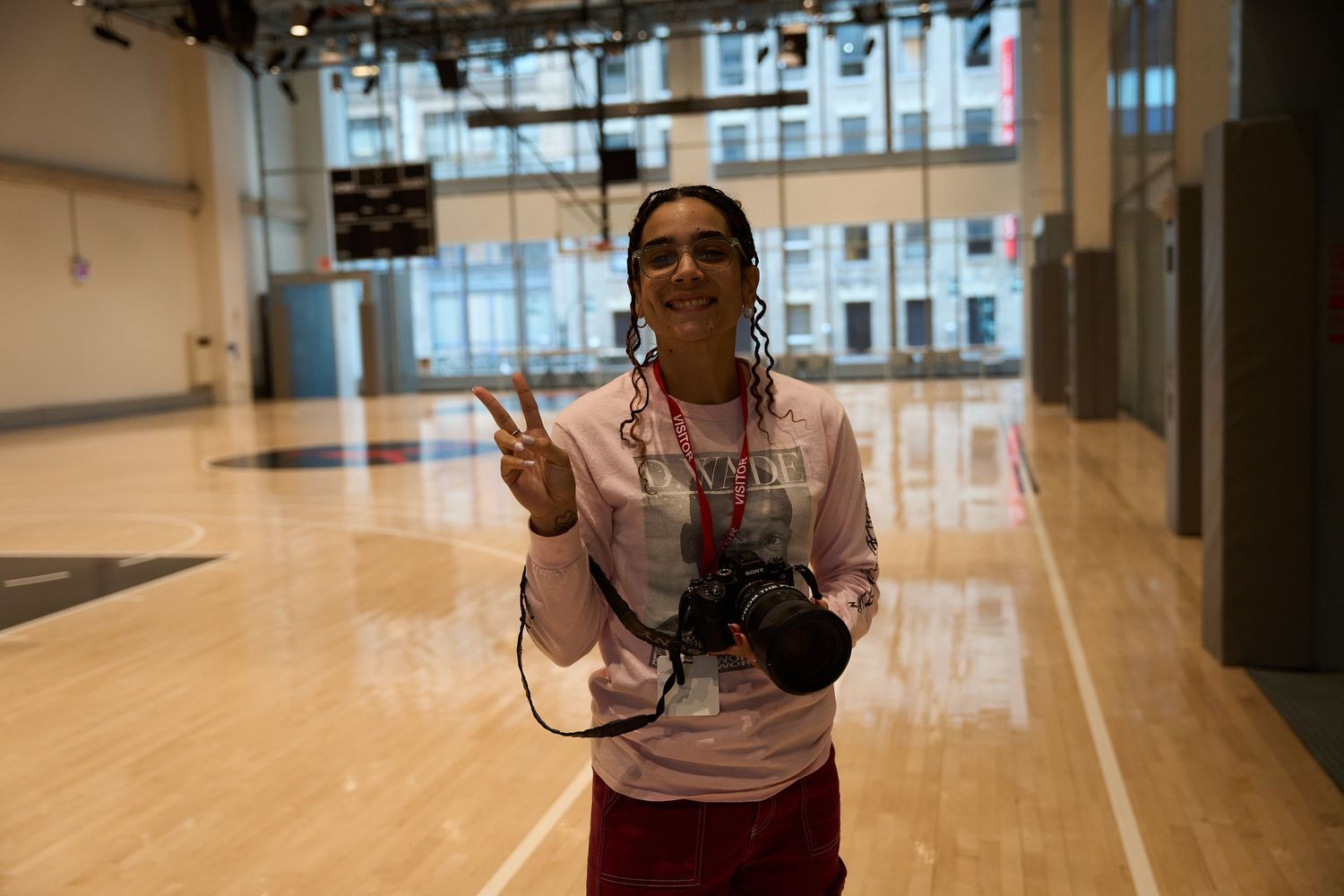 A photographer with a camera flashes a peace sign in an indoor basketball court with polished wooden floors.