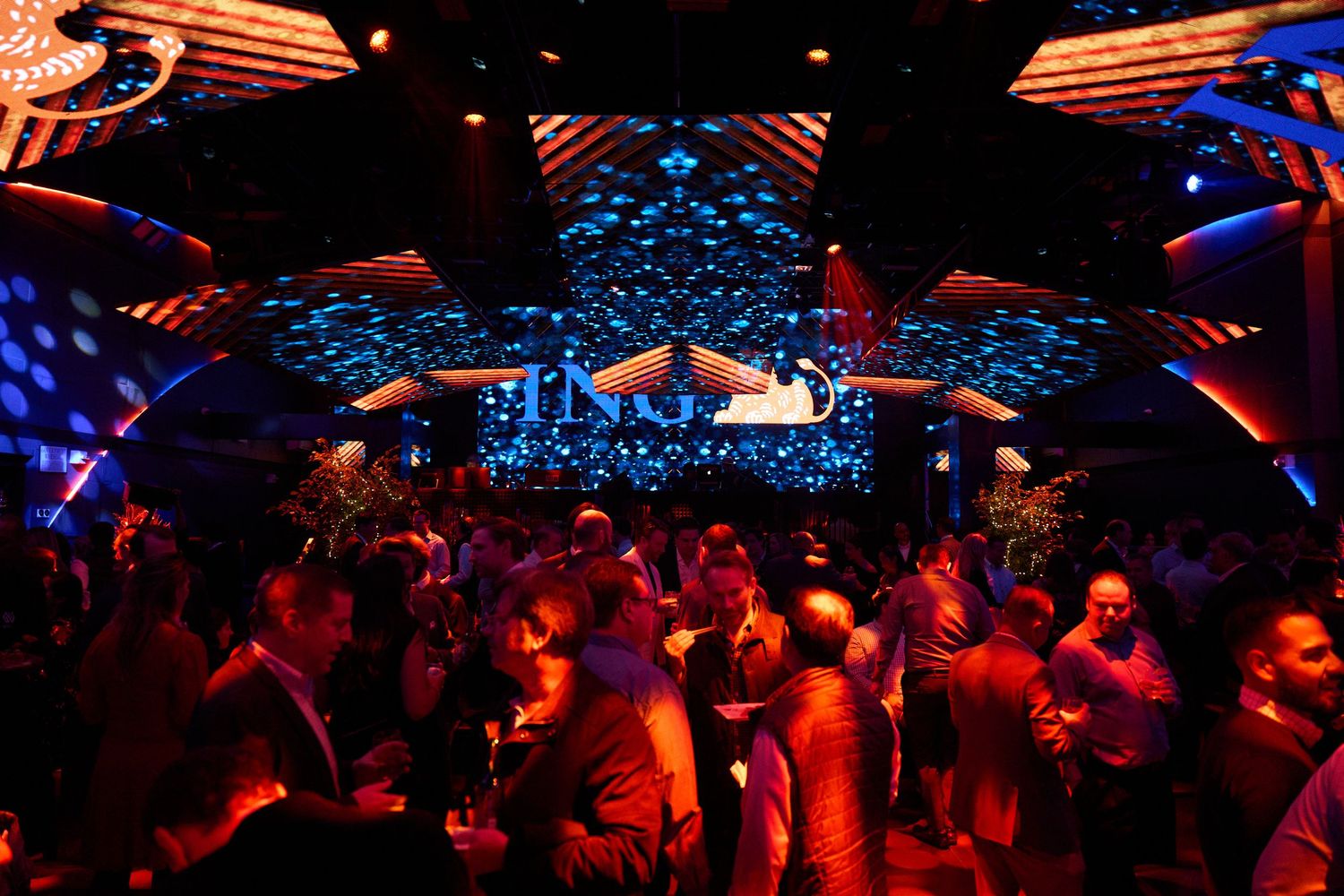 A crowded nightclub scene with vibrant red and blue lighting illuminating the dance floor and decorative ceiling features.