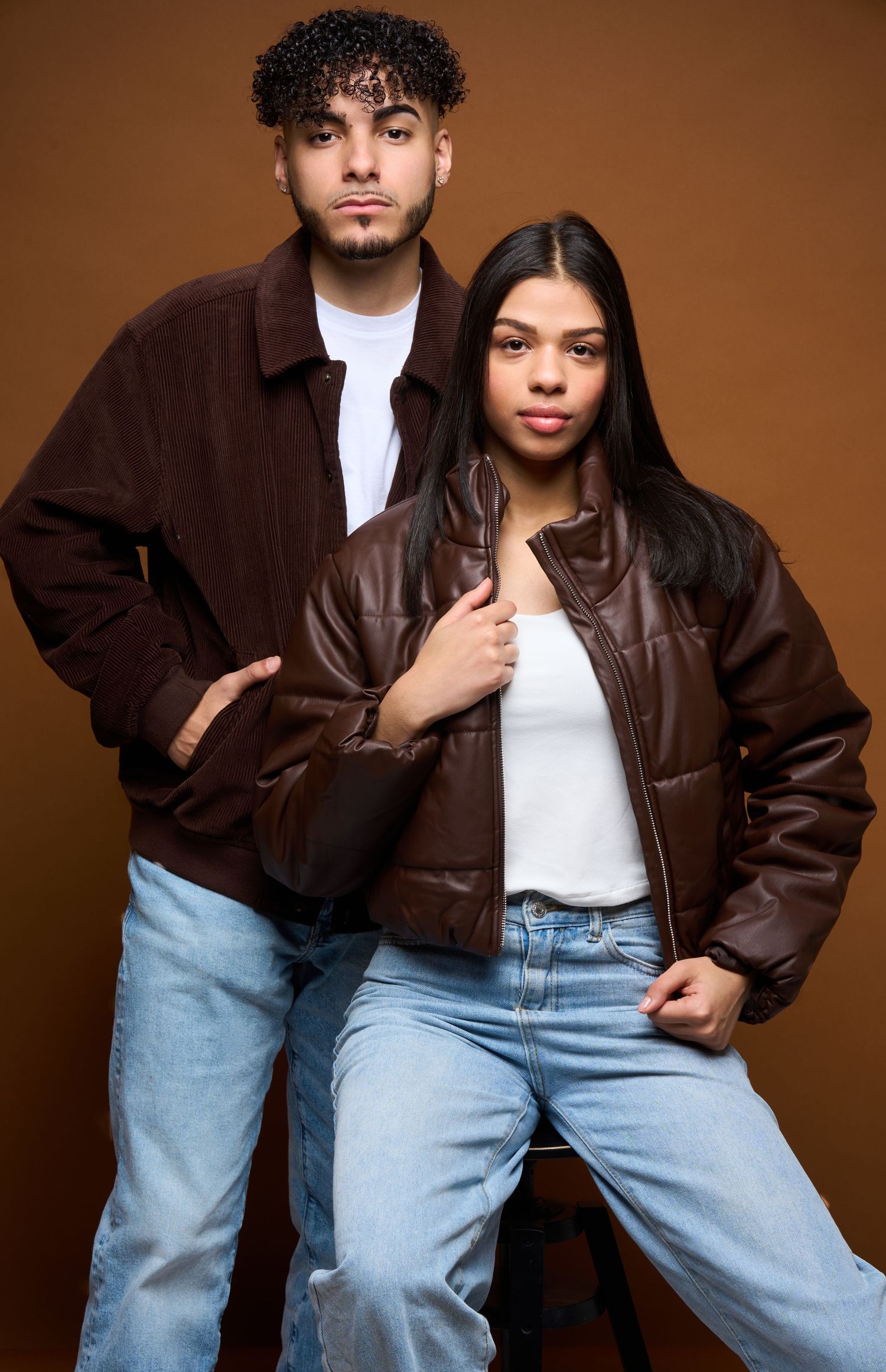 Two people in brown jackets and light jeans pose against a brown backdrop in a casual fashion photoshoot.