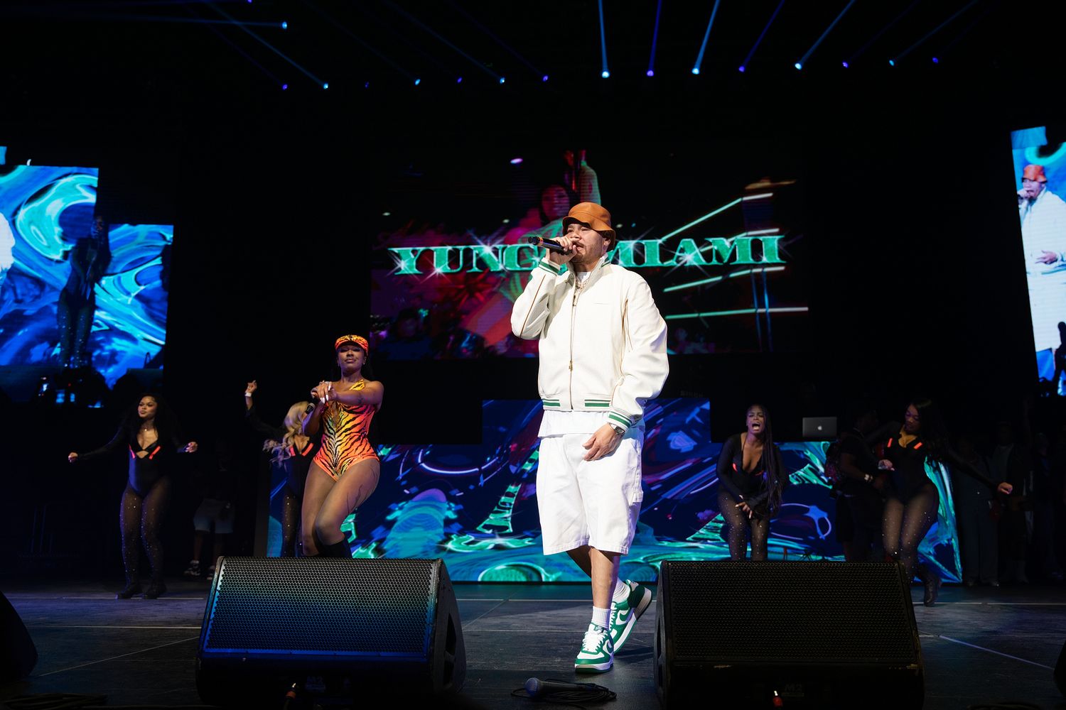 Yung Miami performs live on stage in a white hoodie and shorts with backup dancers and colorful LED screens.