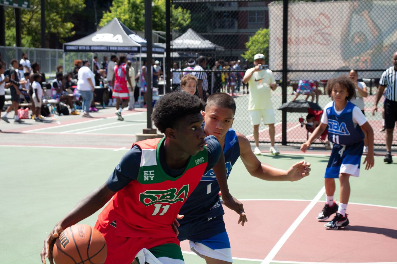 Players competing on an outdoor basketball court during a sunny day with spectators watching from the sidelines.