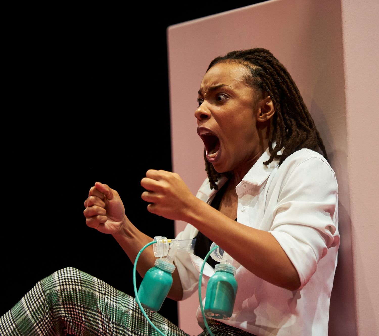 Performer with dreadlocks wearing a white shirt and plaid pants makes a dramatic expression while gesturing on stage.