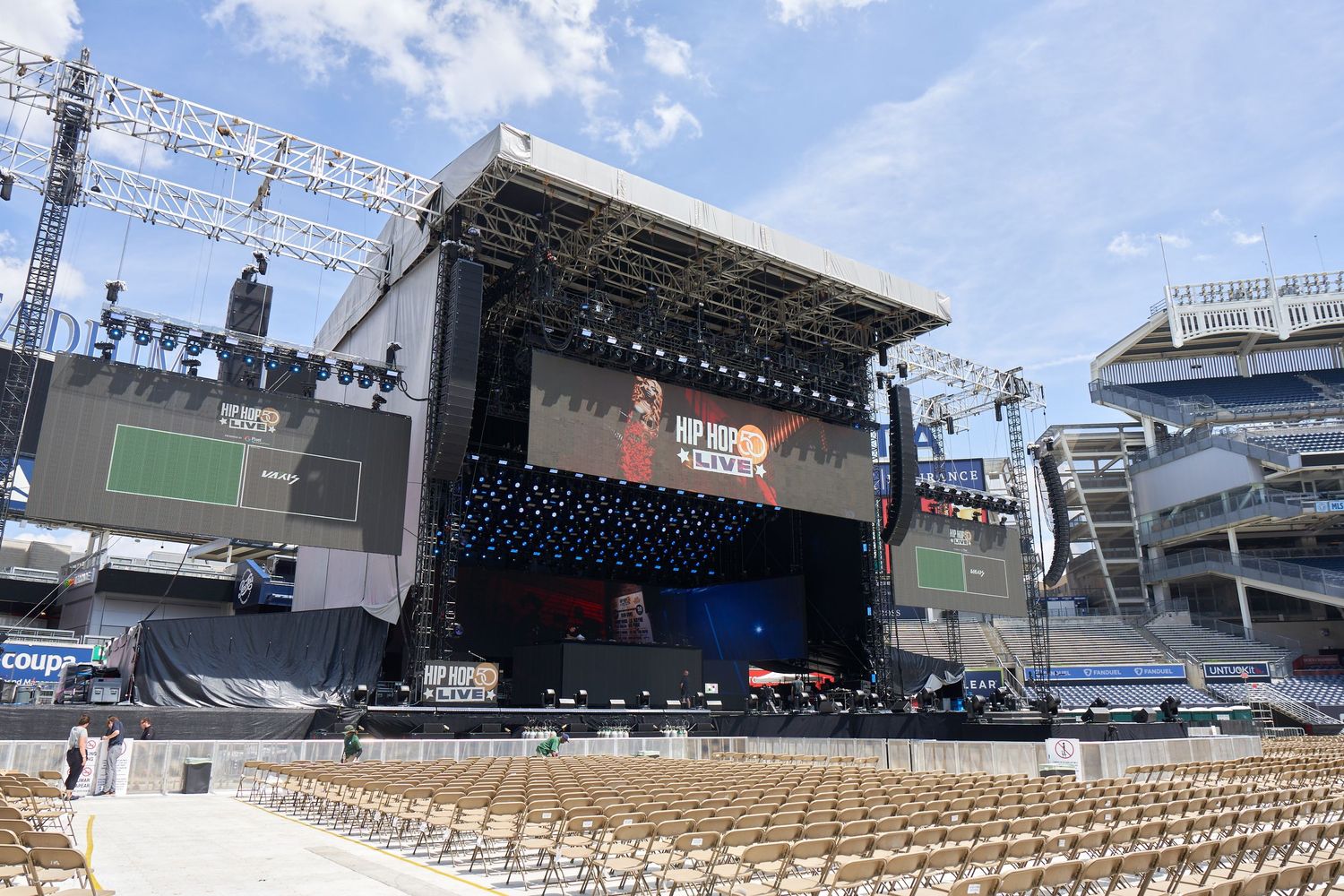 Wide panoramic view of an empty outdoor concert venue with multiple stages and seating areas set up under a blue sky.