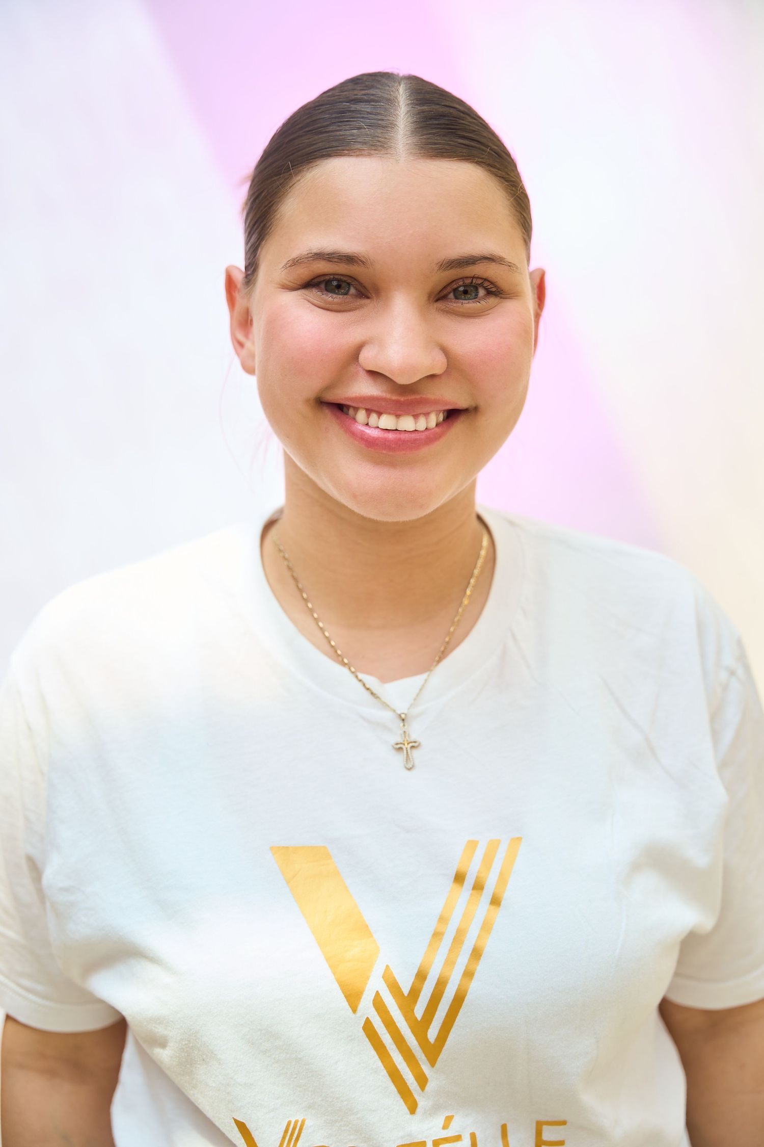Young woman smiling in white V-logo t-shirt with gold cross necklace.