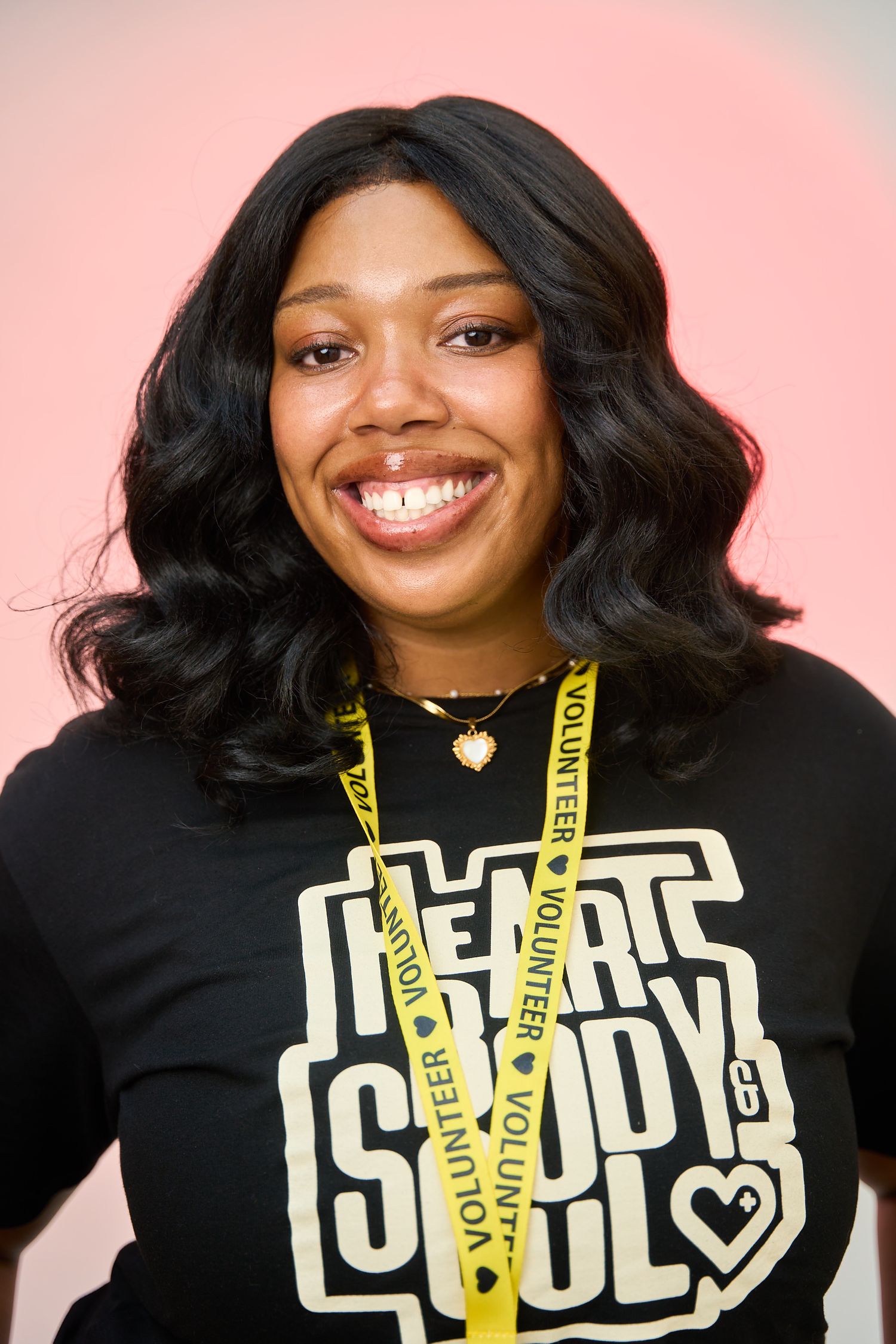 Smiling young woman wearing a black Heart Body Soul t-shirt and yellow volunteer lanyard against a pink background.