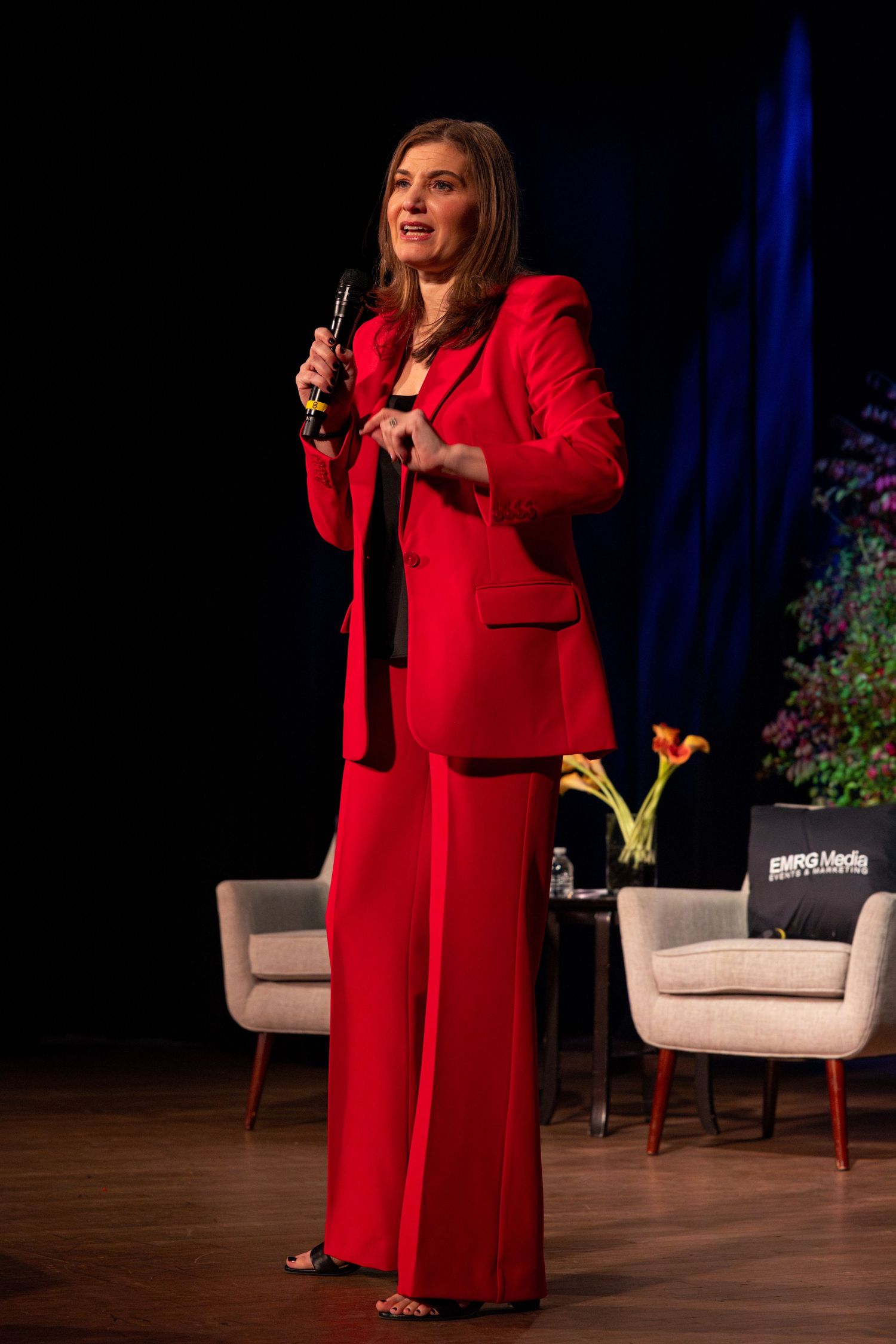 A speaker in a bright red coat gestures while presenting on a dark stage with white chairs and floral arrangements.