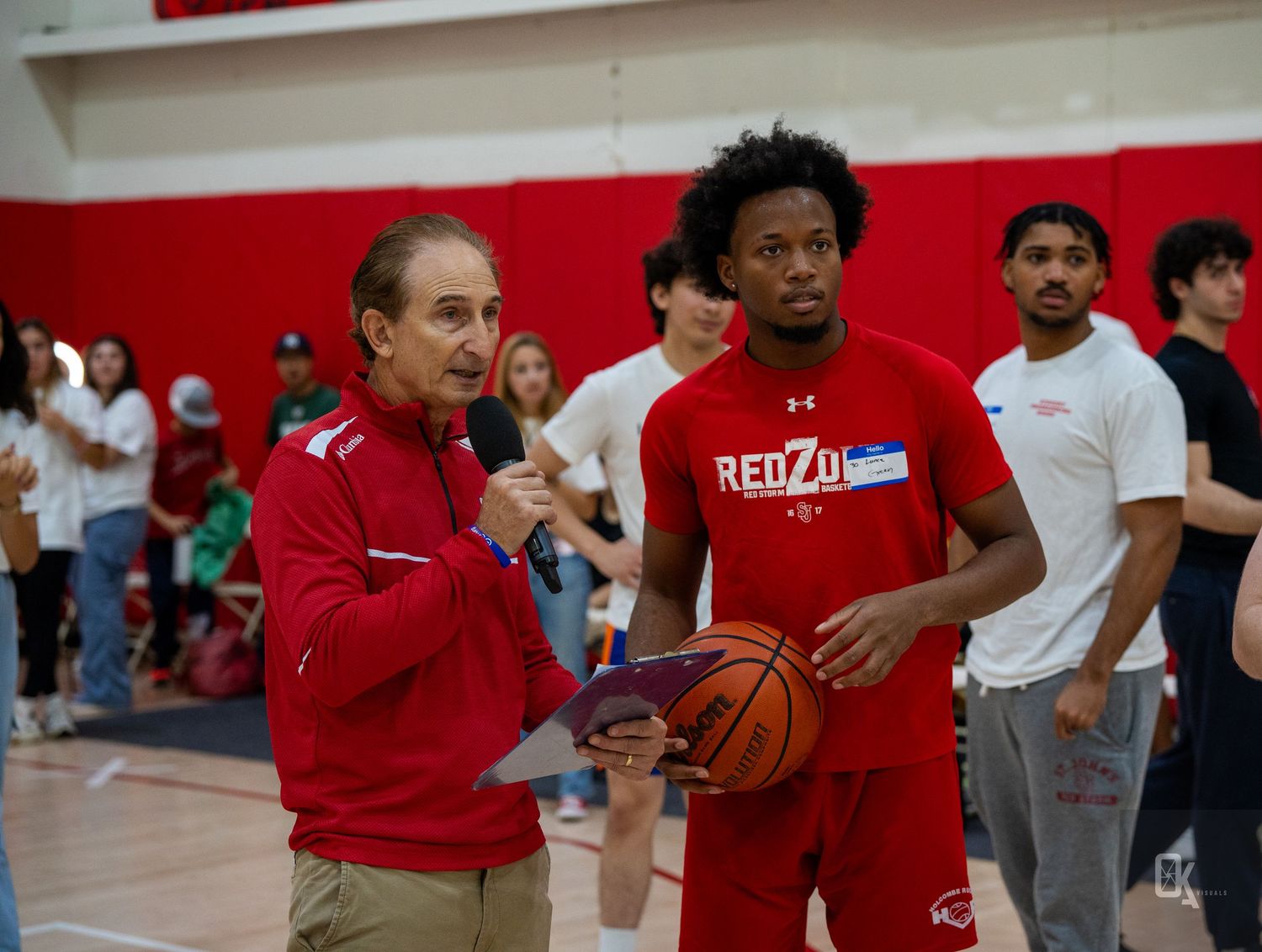 A basketball coach in red attire provides instruction during practice in a gymnasium with red and white walls.