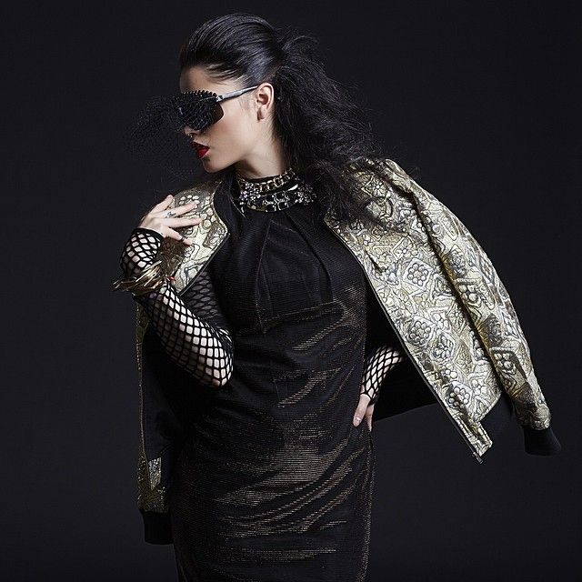 Fashion model wearing black outfit with snakeskin print jacket poses dramatically against dark background.