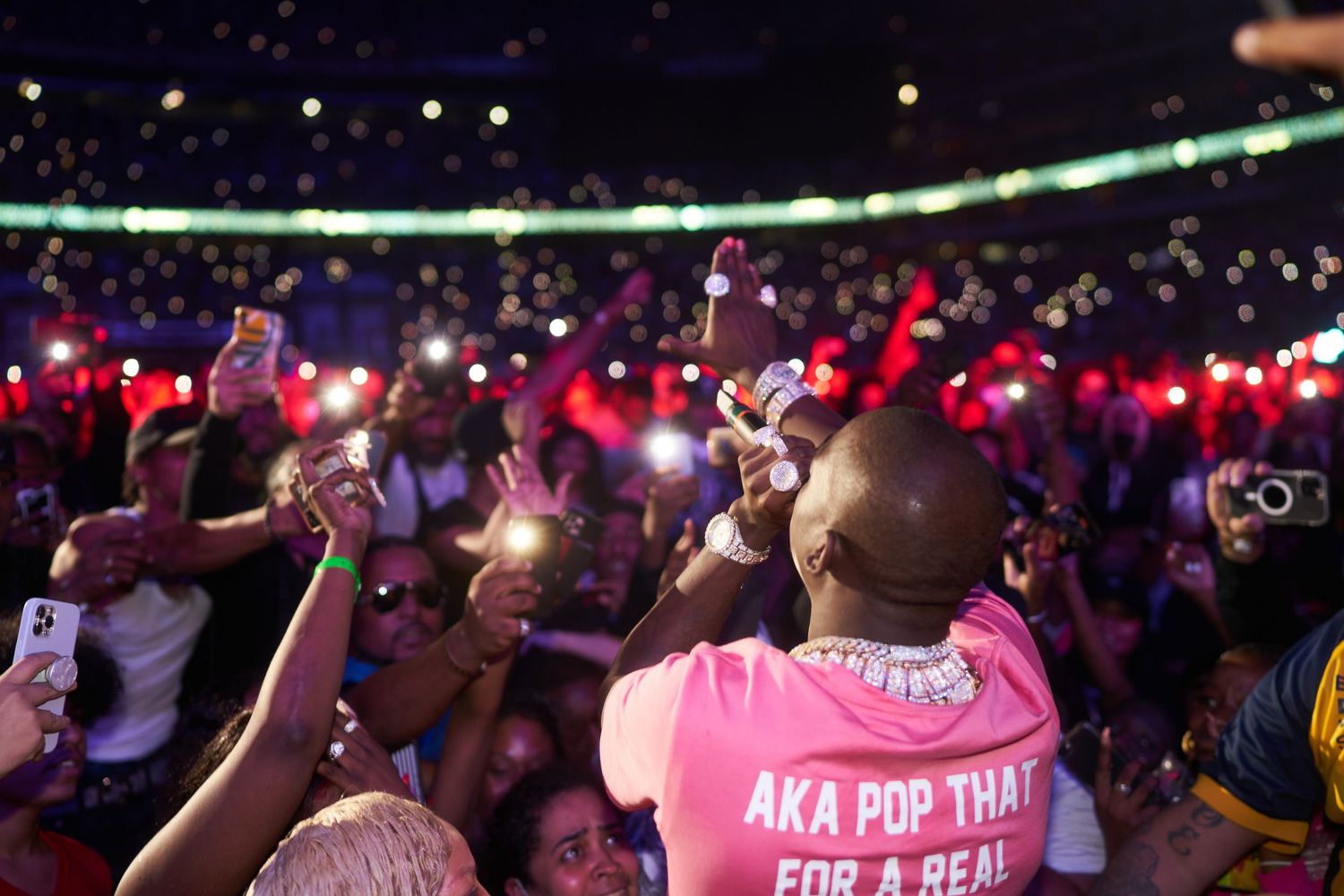 Concert crowd holds up phones with glowing red lights while someone in a pink t-shirt captures the moment.