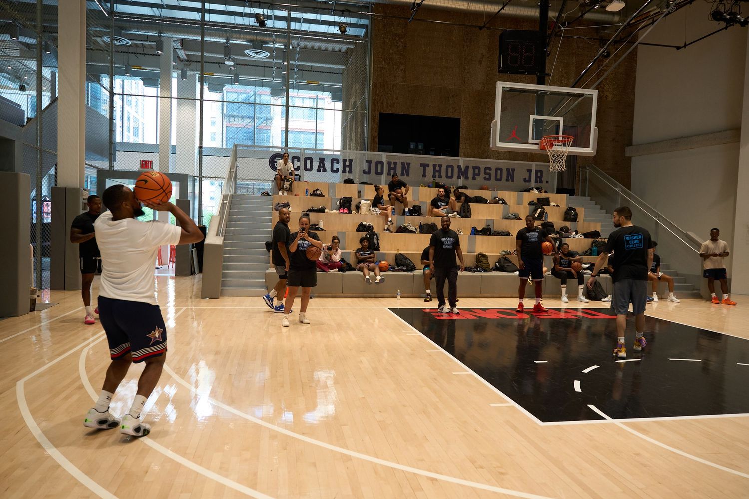 Indoor basketball court with players practicing and running drills in a modern facility with natural lighting.