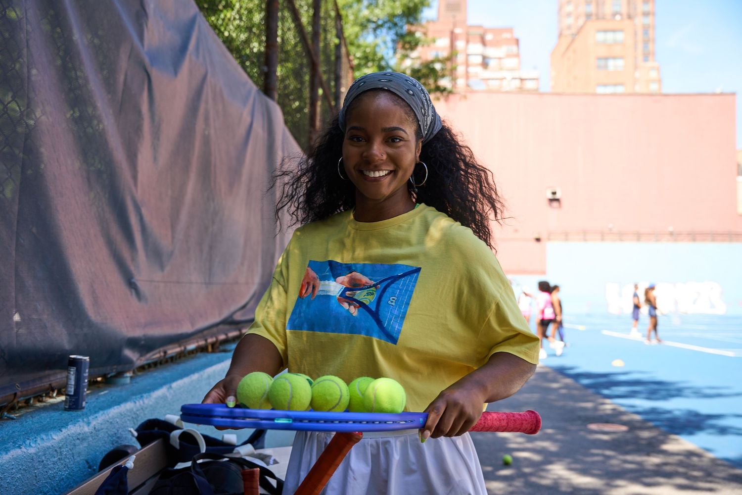 A tennis ball collector stands with a basket of tennis balls on a sunny court.