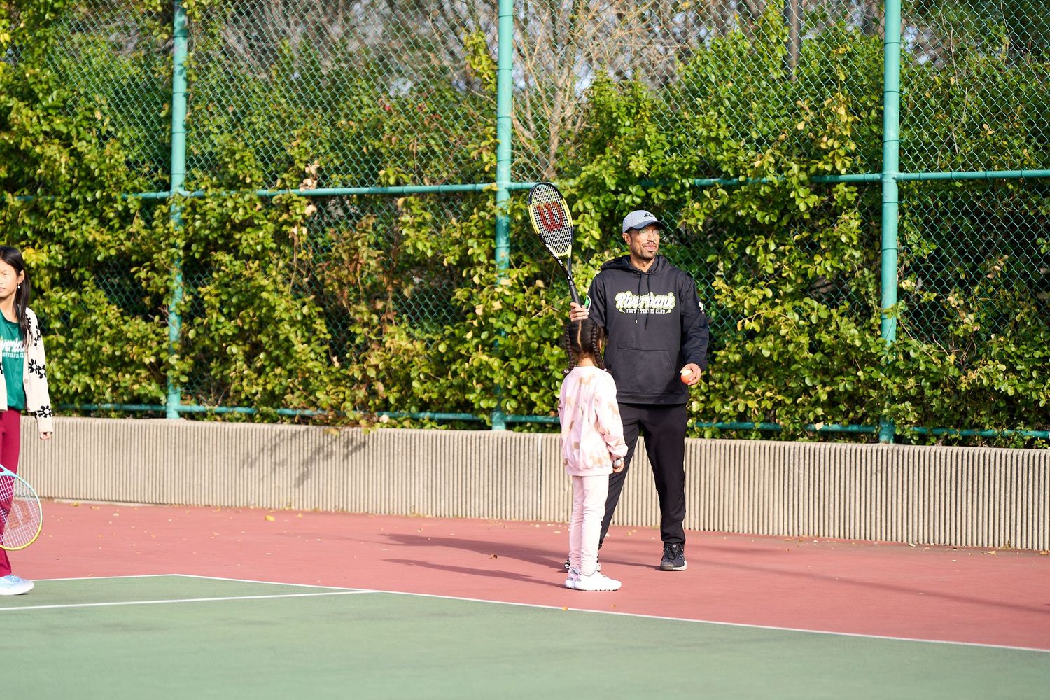 People playing tennis on an outdoor red court with green fencing and bushes in the background.