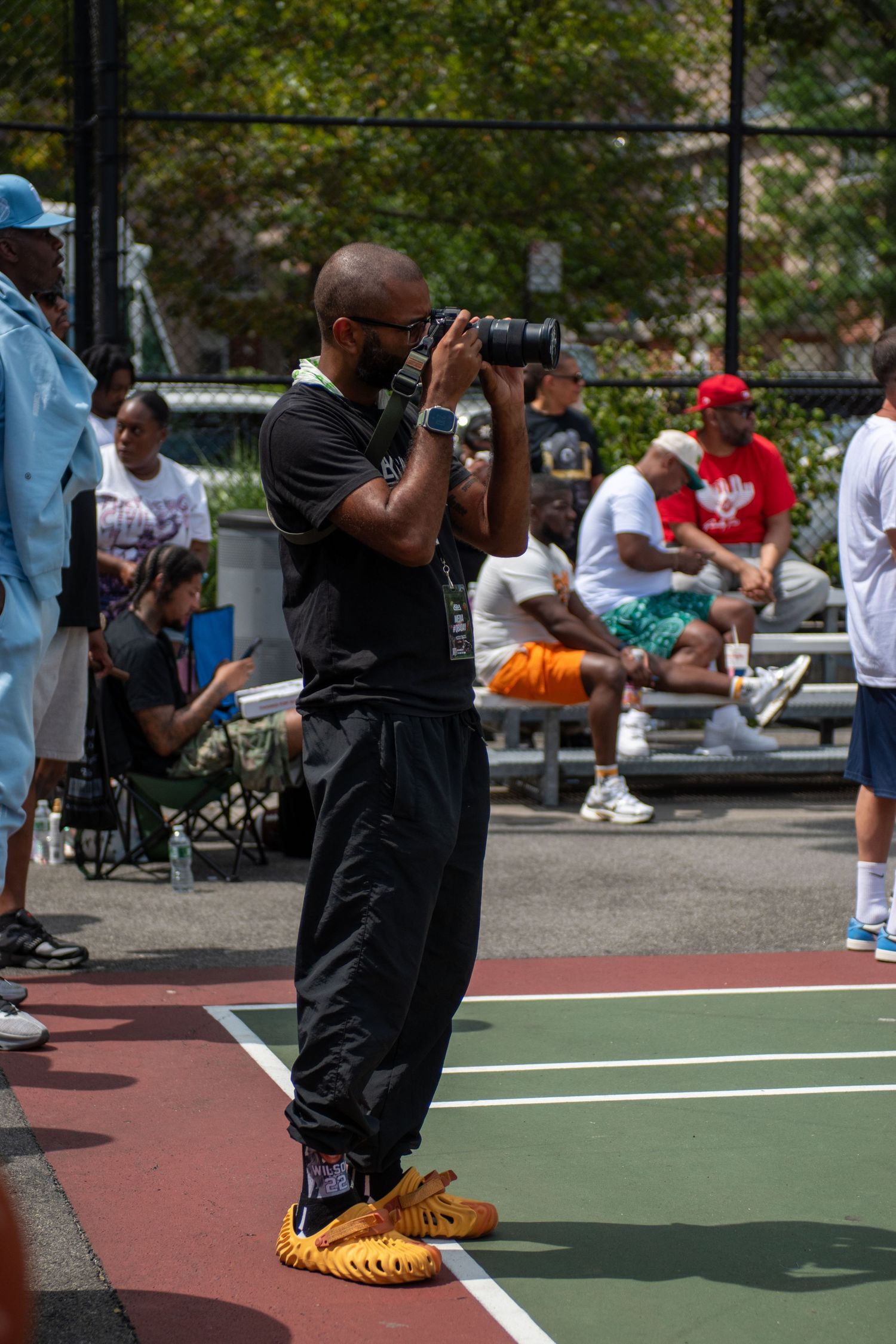 A photographer wearing black clothes and yellow shoes takes pictures at an outdoor basketball court with spectators in background.