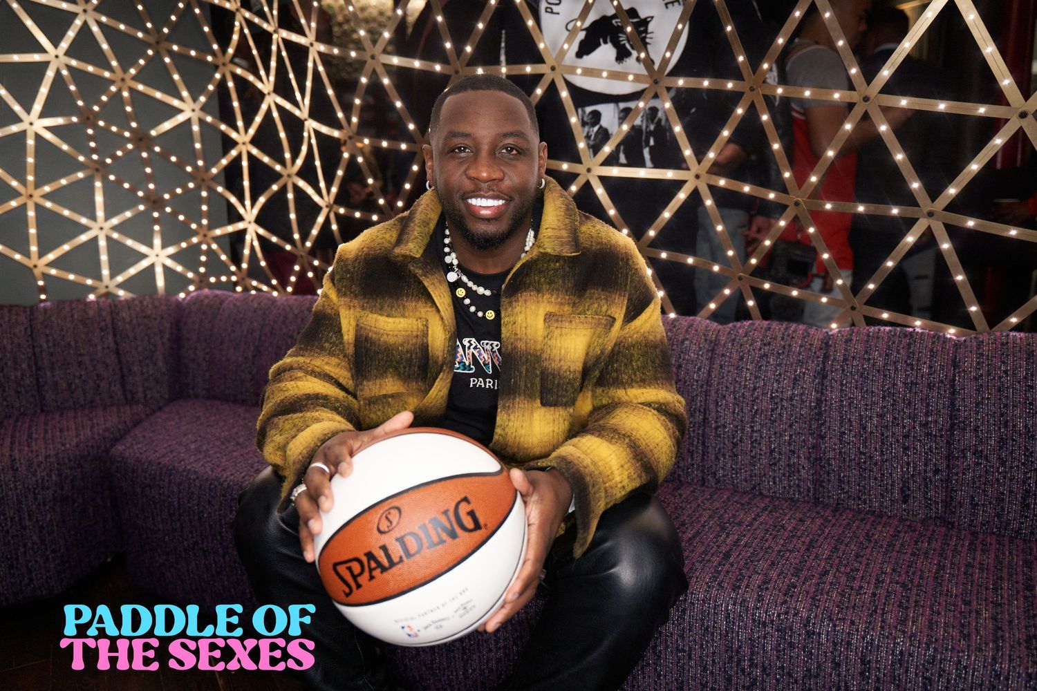 Person wearing yellow plaid jacket sits on purple couch holding basketball with decorative light display background.