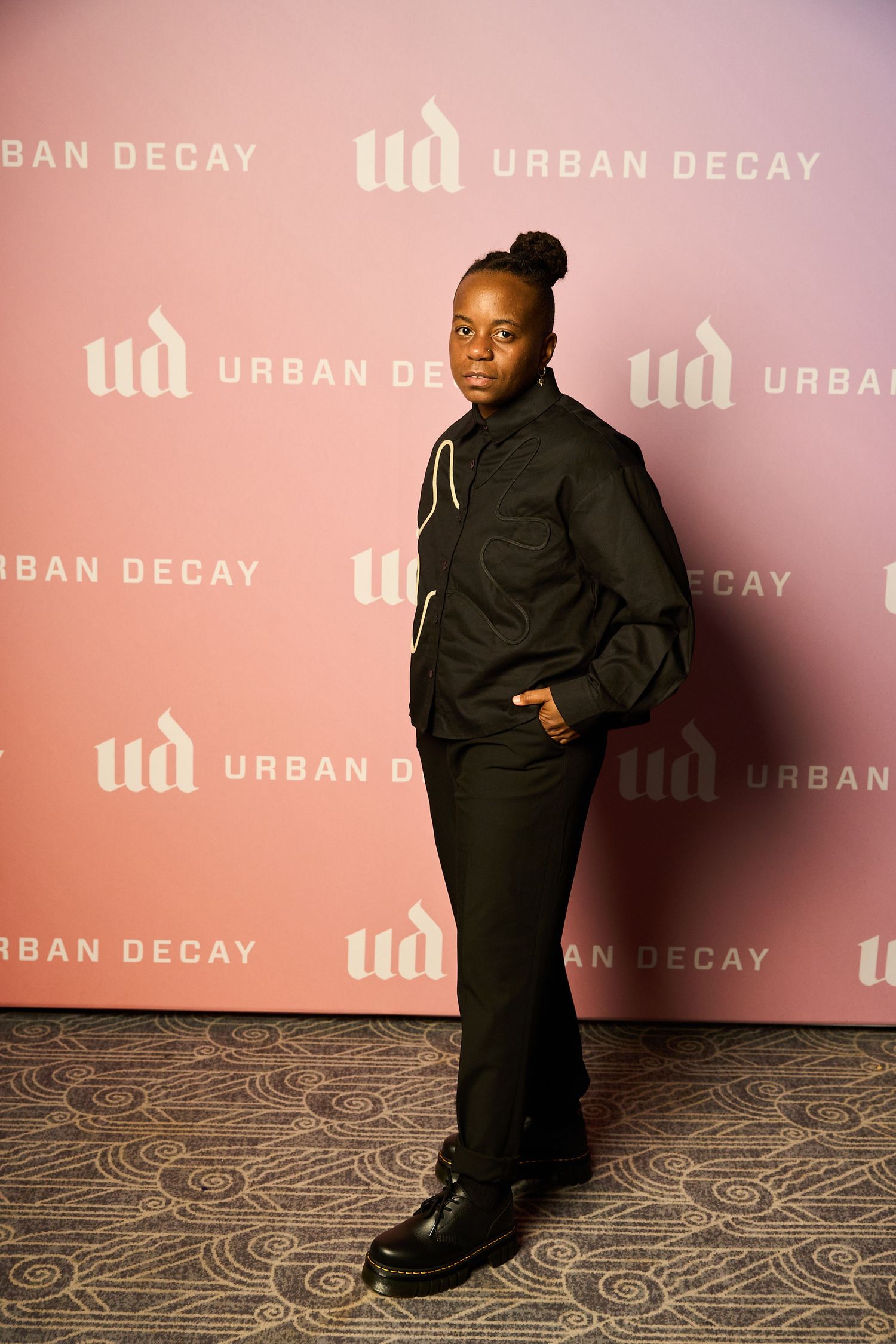 A figure in black clothing poses against a pink Urban Decay branded backdrop while seated on the floor.