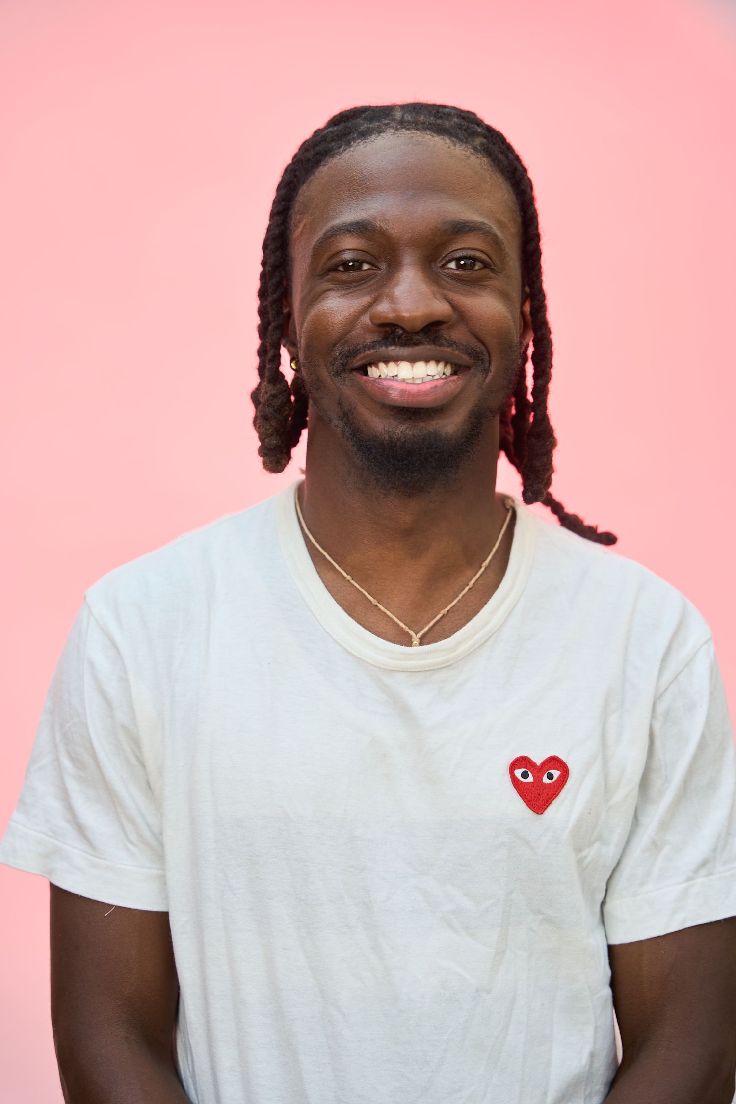 Smiling young man with braids wearing a white Comme des Garçons t-shirt against a pink background.