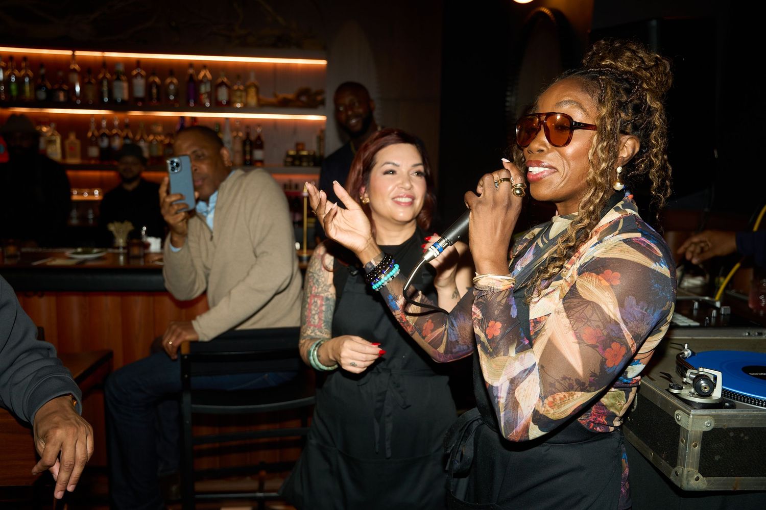 Woman in sunglasses and a colorful patterned jacket sings into a microphone at a lively bar while friends smile and laugh nearby.