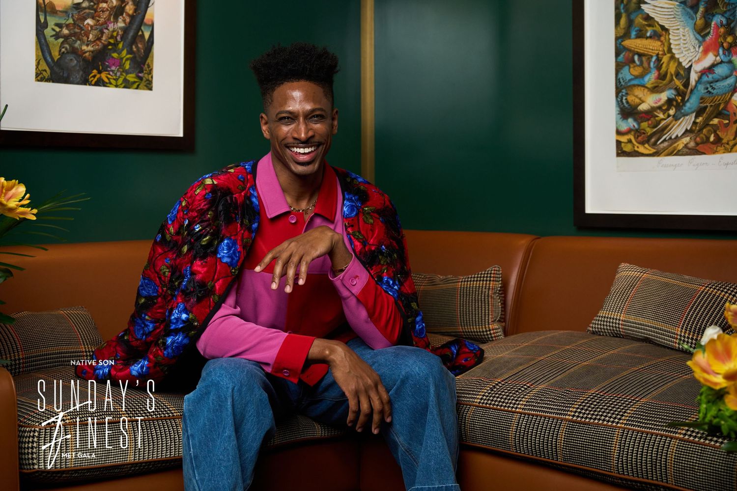 A person sits on a plaid couch wearing a colorful patterned shirt and jeans against a green wall with framed art.