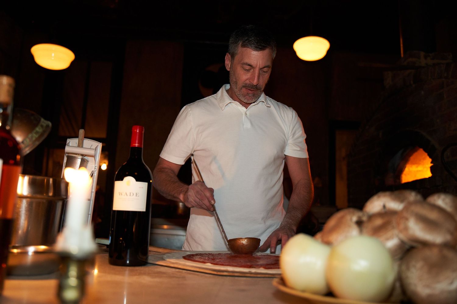 Chef in white polo shirt preparing food at dimly lit restaurant bar with wine bottles and onions visible.