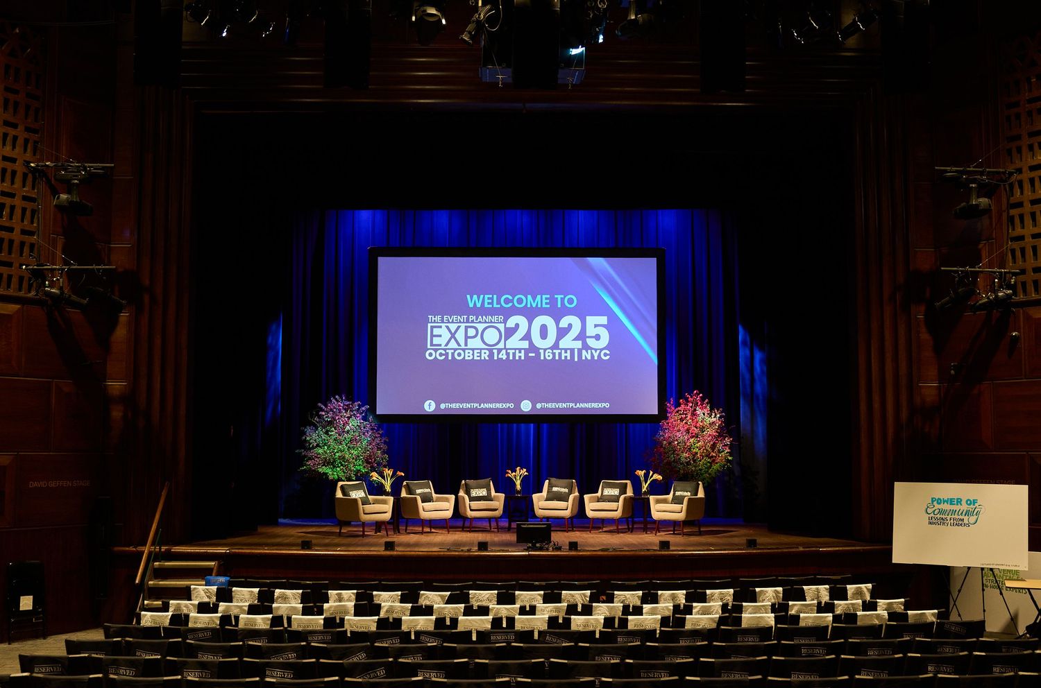Theater stage with blue-lit screen displaying 'PAX 2025' surrounded by floral arrangements and empty auditorium seats.