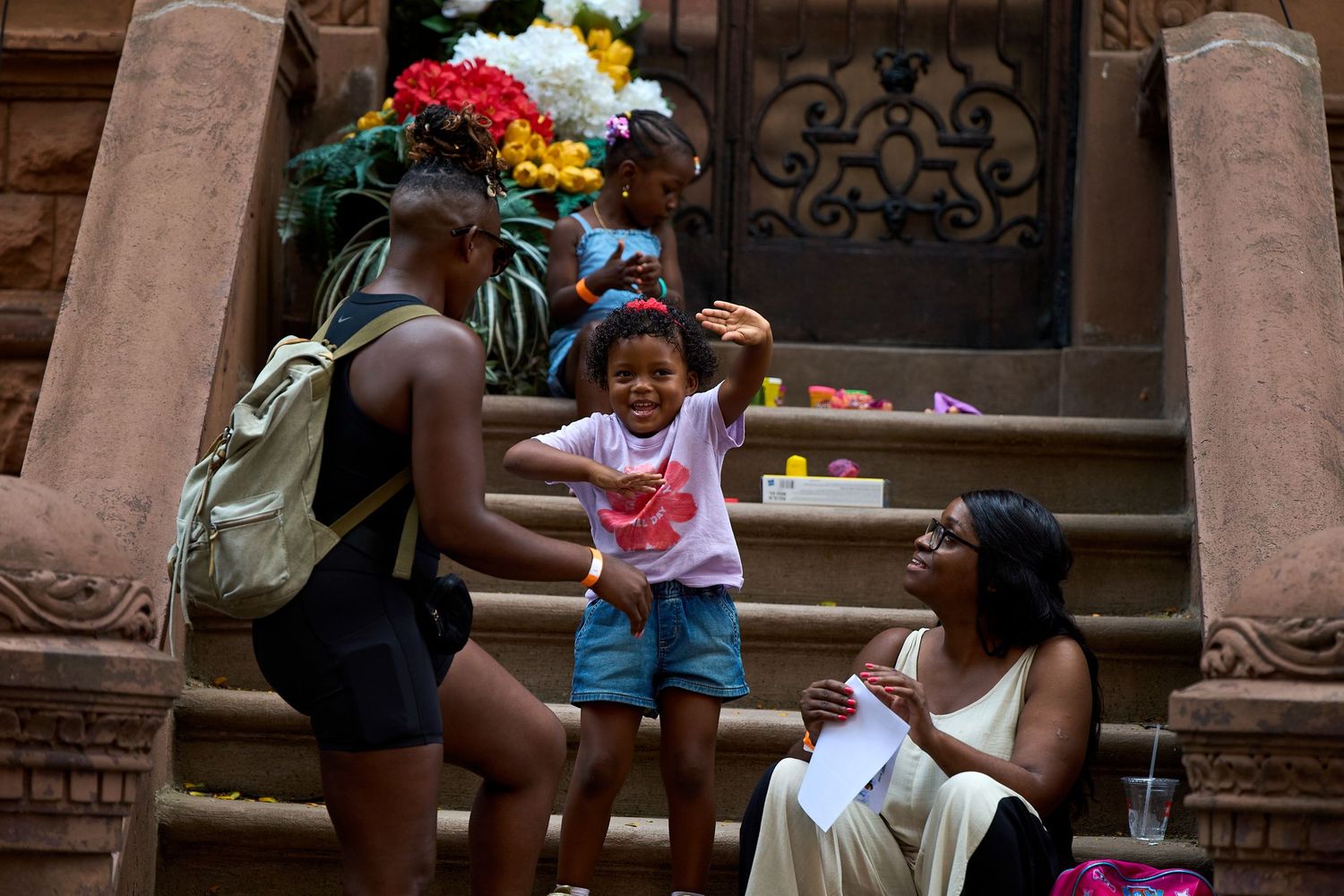 A child in a pink shirt and blue shorts stands joyfully on brownstone steps while others sit nearby.