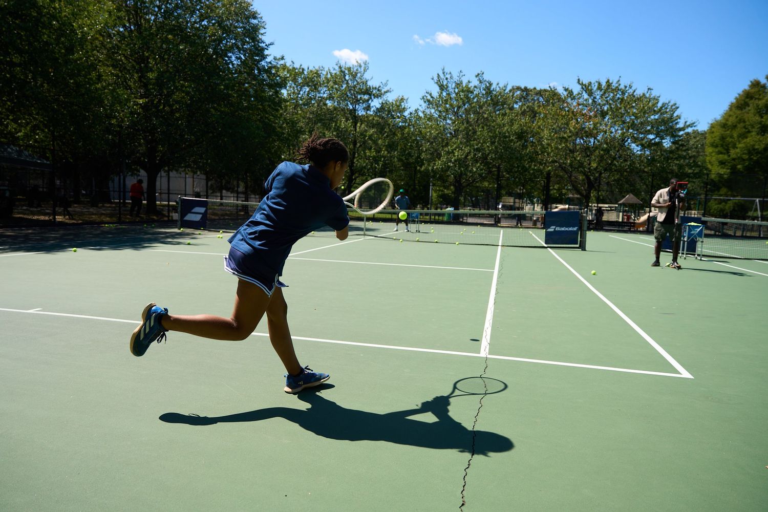 Multiple players practicing tennis drills on a green outdoor court with trees in the background.