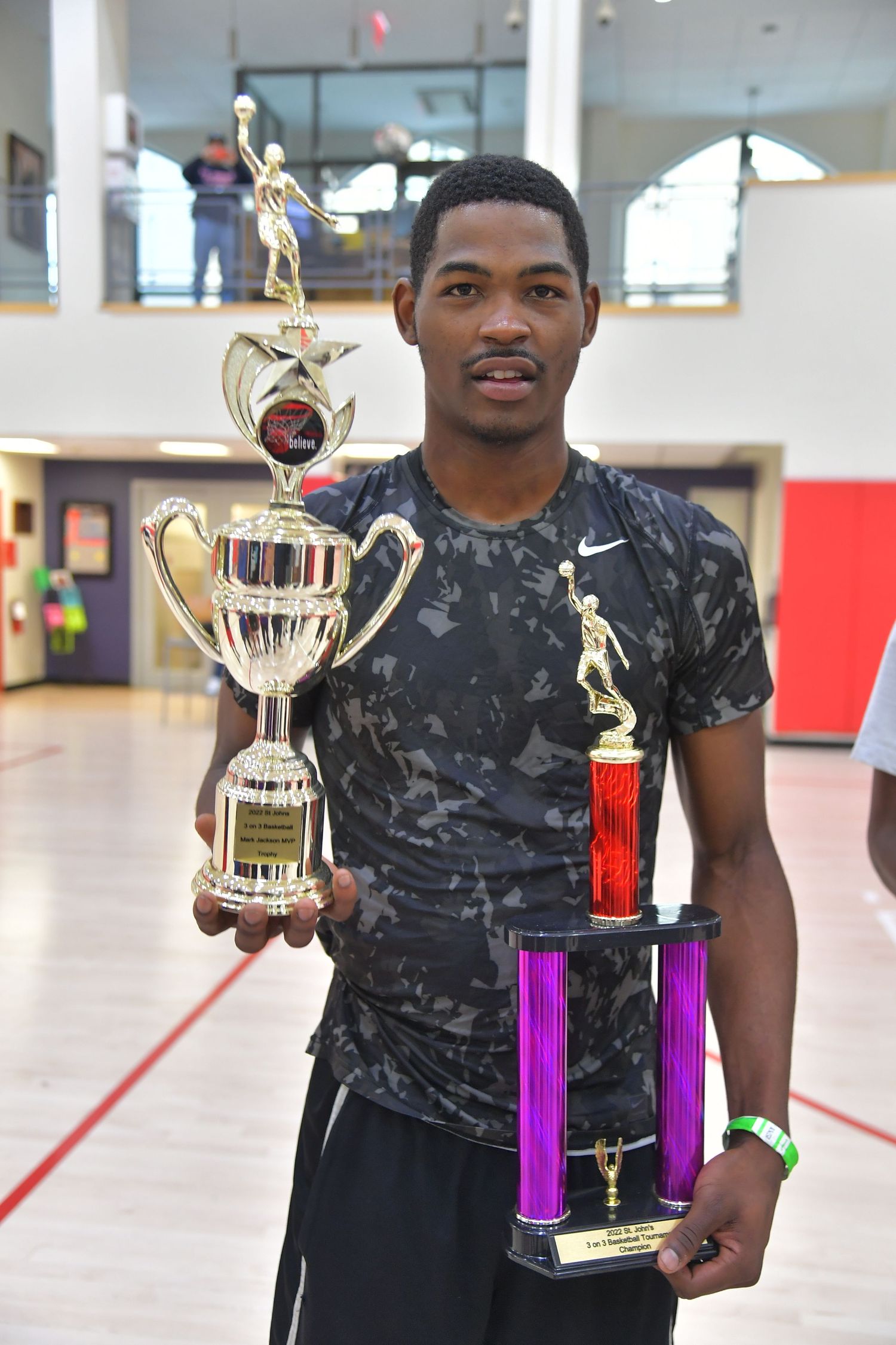 Person in black camo shirt holds gold trophy awards in indoor gym setting.