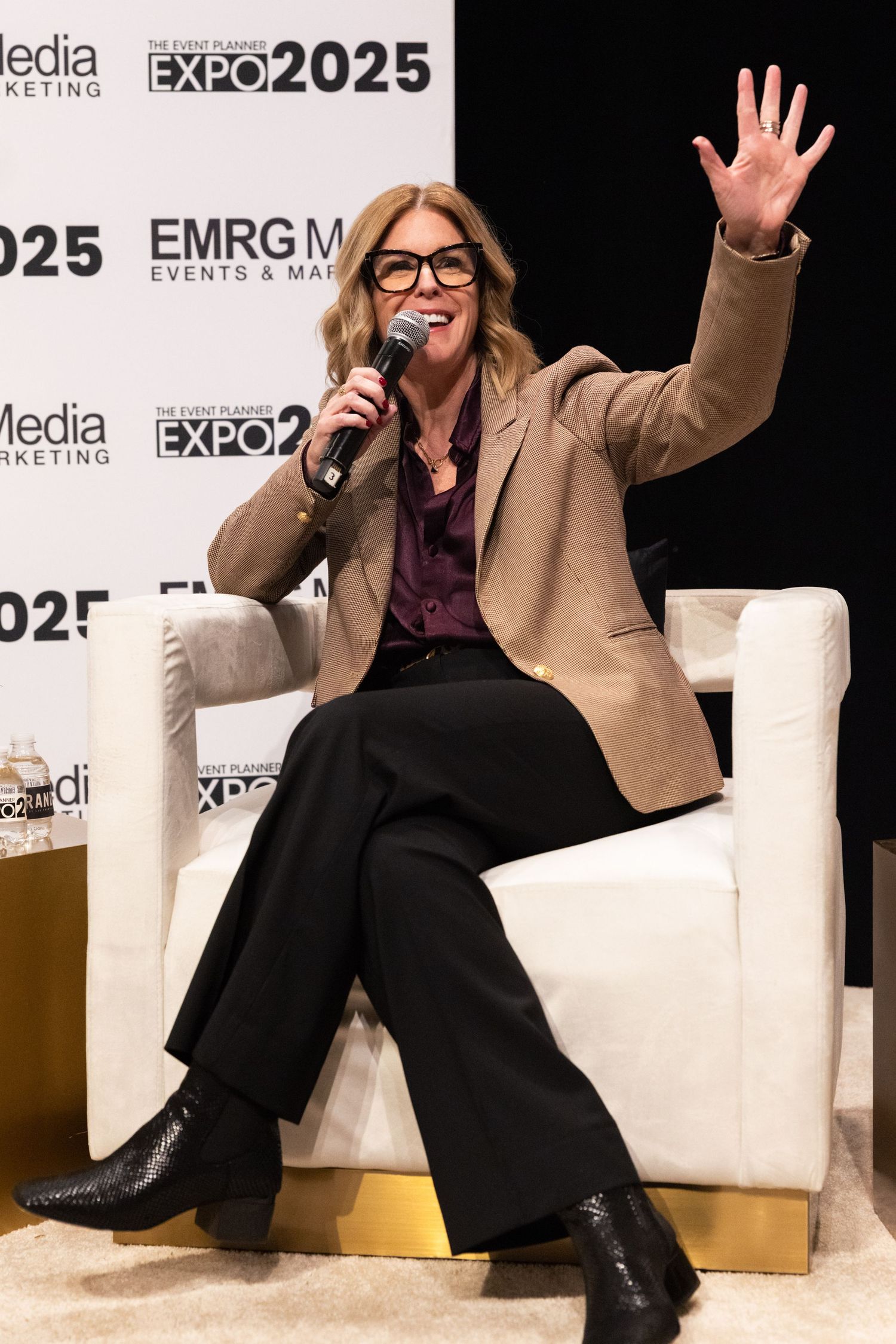 Speaker in tan blazer and glasses gestures animatedly while seated on white chair at Expo 2025 event.