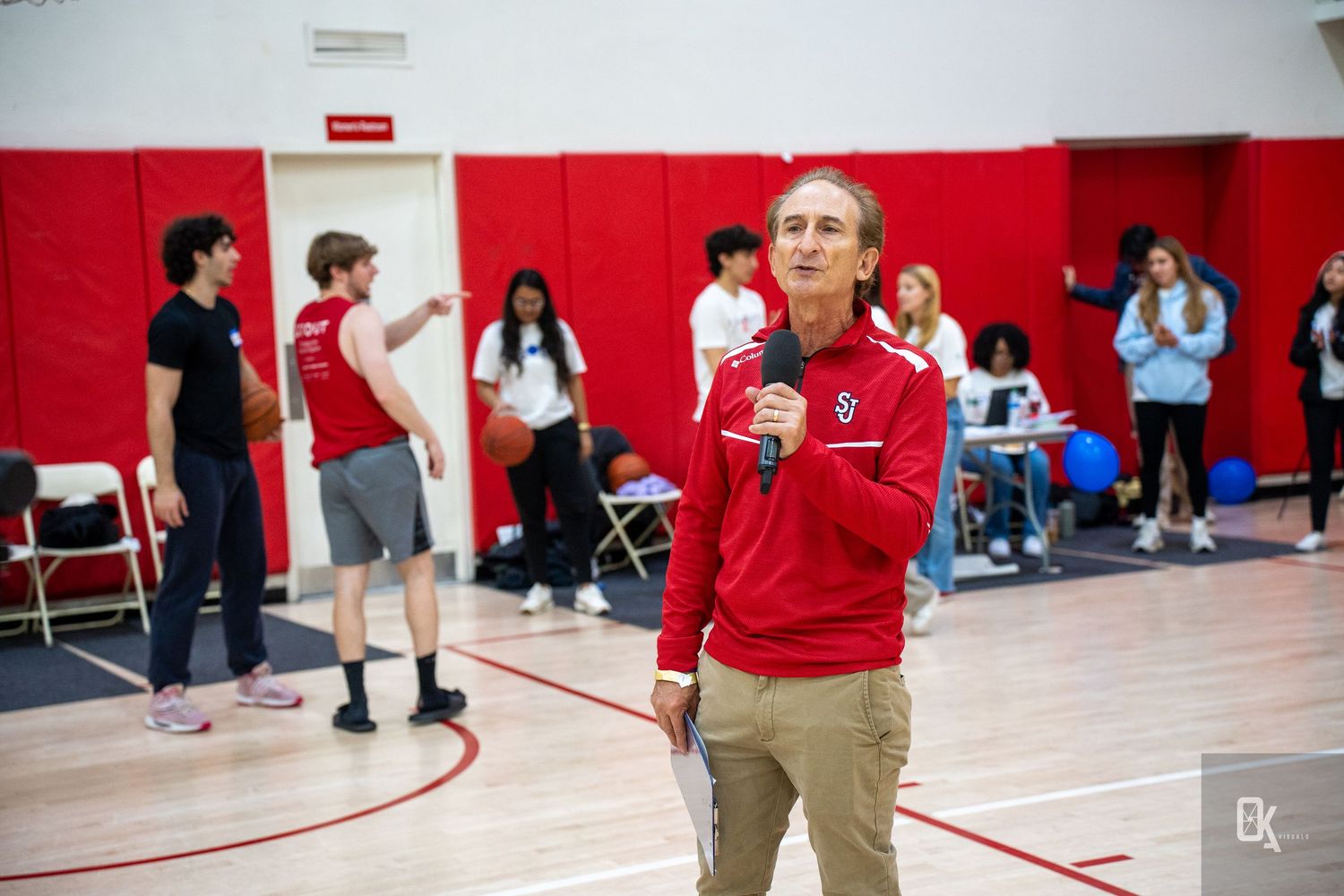 Basketball players and spectators gather in a red-walled gymnasium during a sporting event.