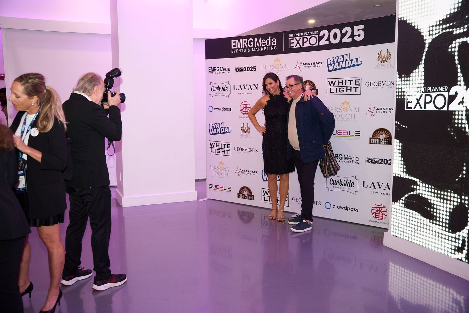 Event photo backdrop with sponsor logos and purple lighting at an expo venue.