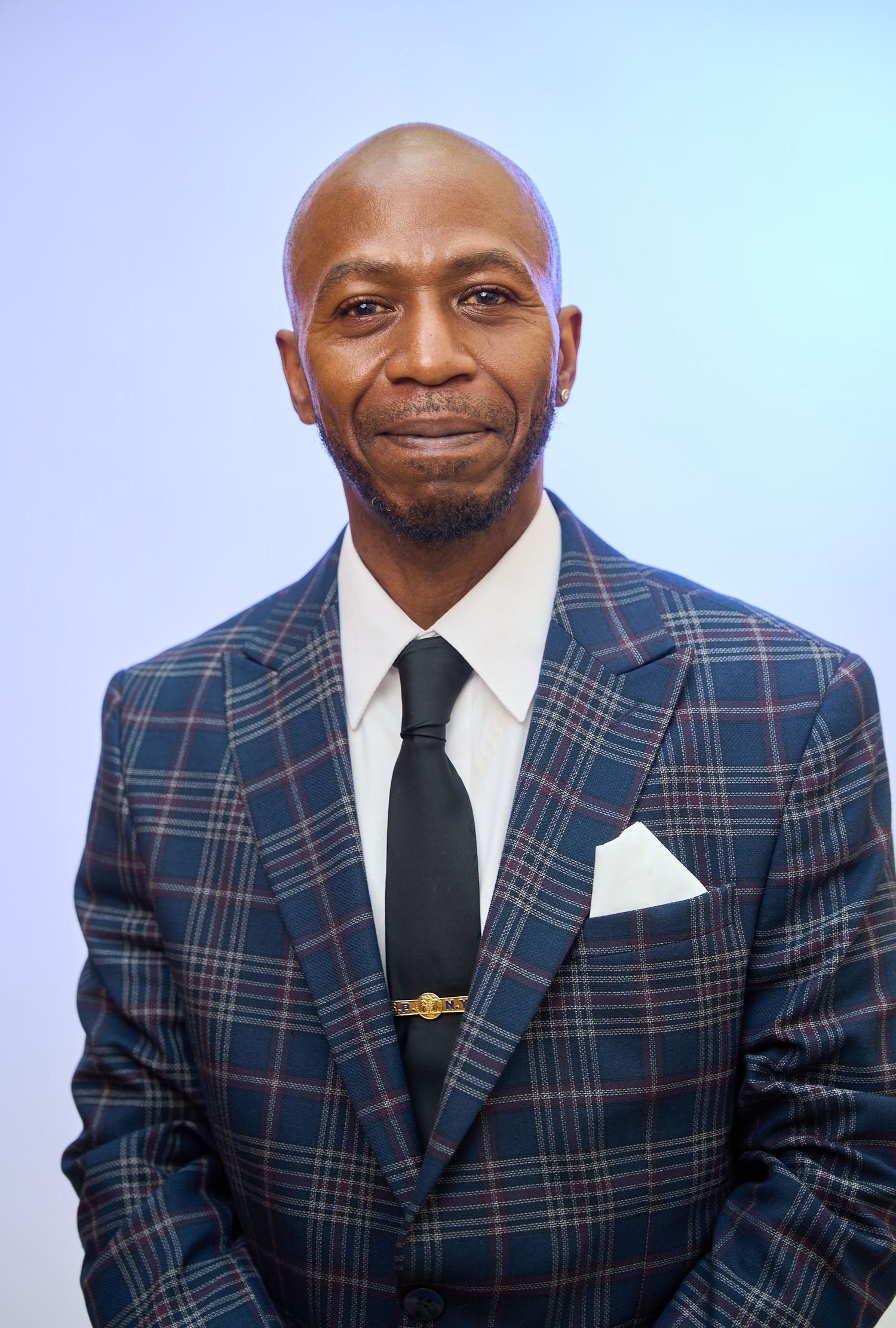 Bald man in plaid blazer with black tie and white pocket square poses confidently.