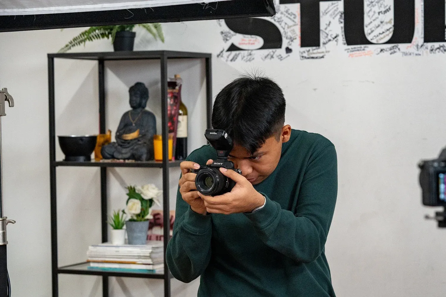 A photographer in a dark green sweater takes a photo while a decorative shelf with Buddha statue is visible in background.