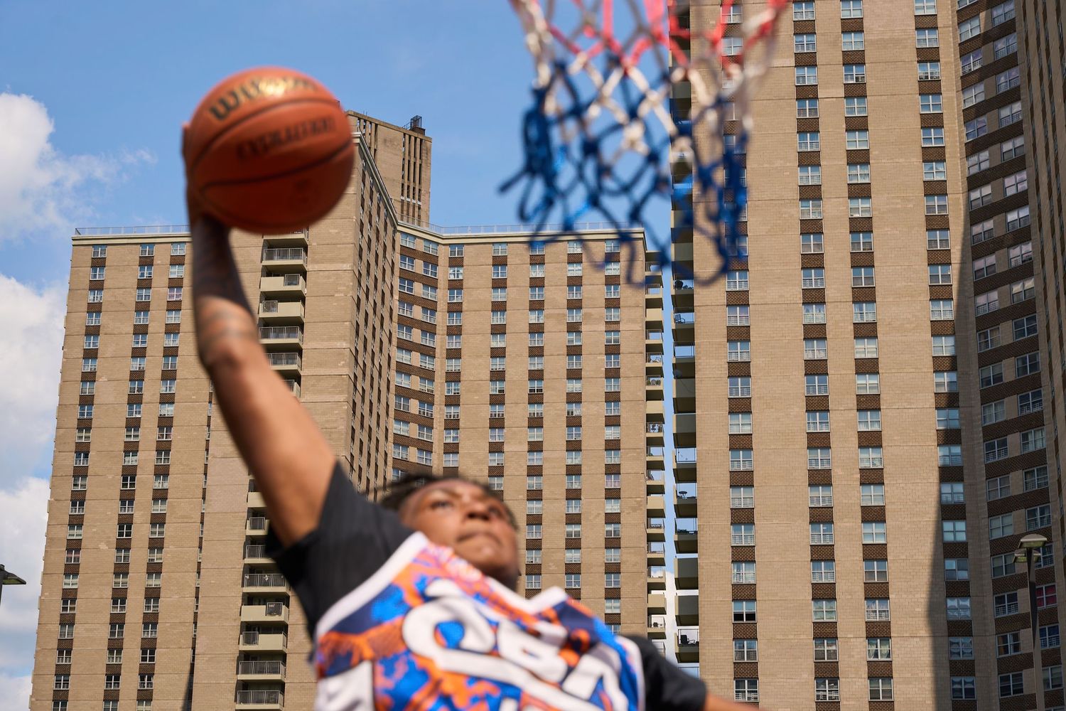 Basketball player wearing red white and blue jersey performs a layup against backdrop of tall apartment buildings.