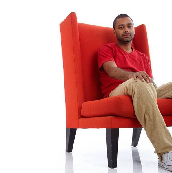A person wearing khaki pants and a red shirt sits casually in a modern red armchair on a white background.