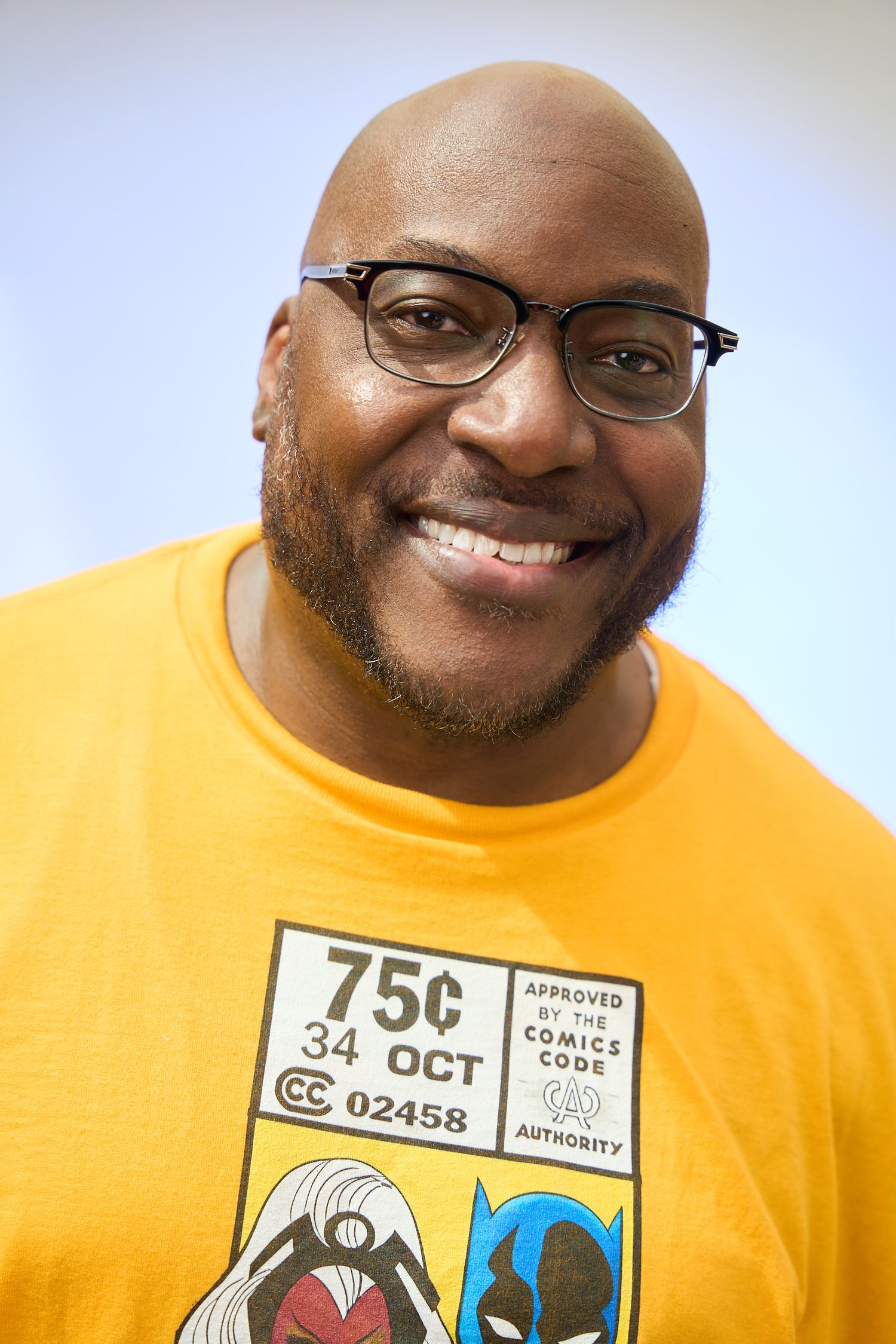 Smiling bald Black man with glasses wearing a yellow comic book themed t-shirt.
