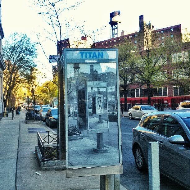Titan payphone kiosk on a city sidewalk at sunset, with parked cars, bare trees and red brick buildings lining the street behind it.