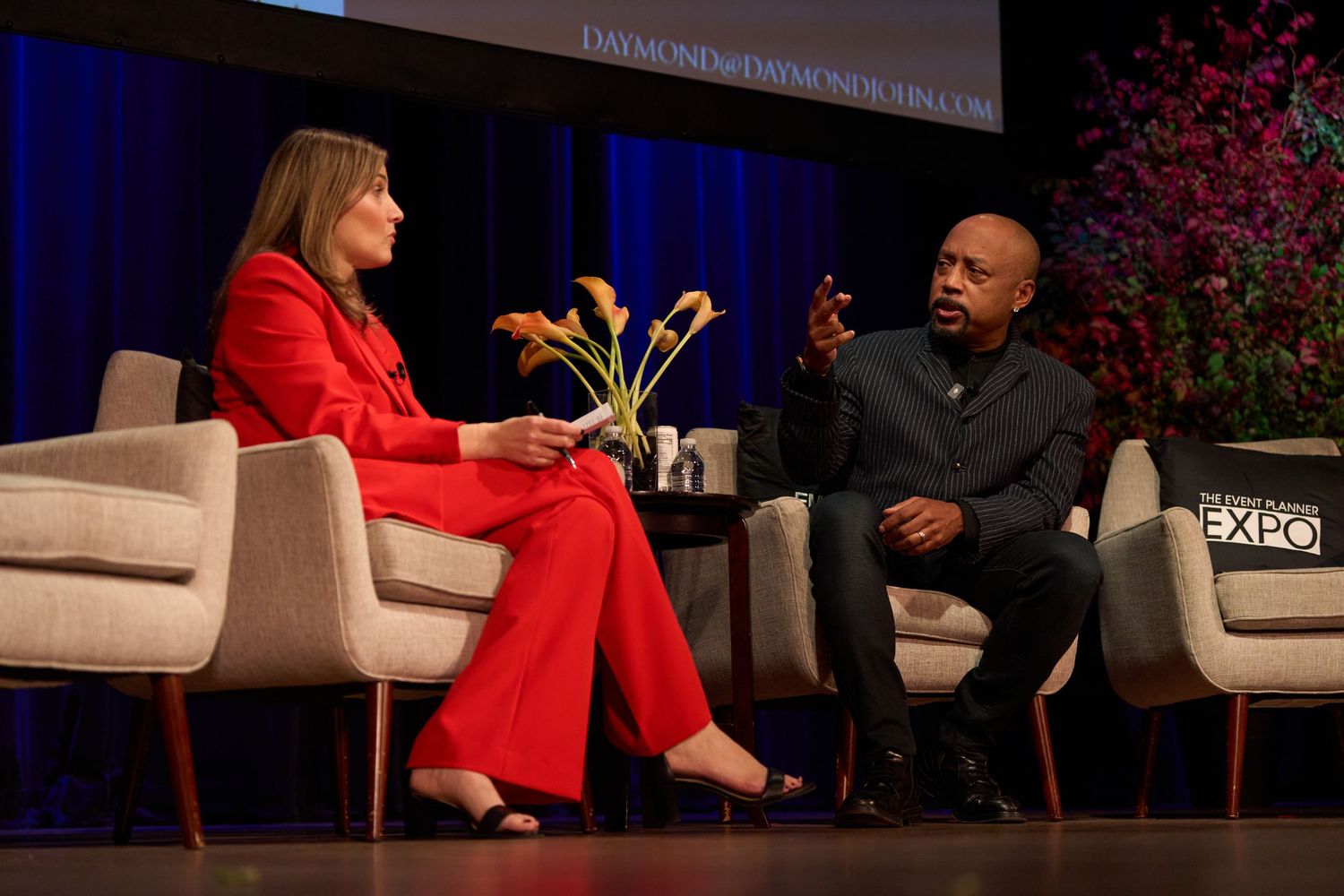 Professional speakers seated in white armchairs having an animated discussion on dimly lit stage with red decor.