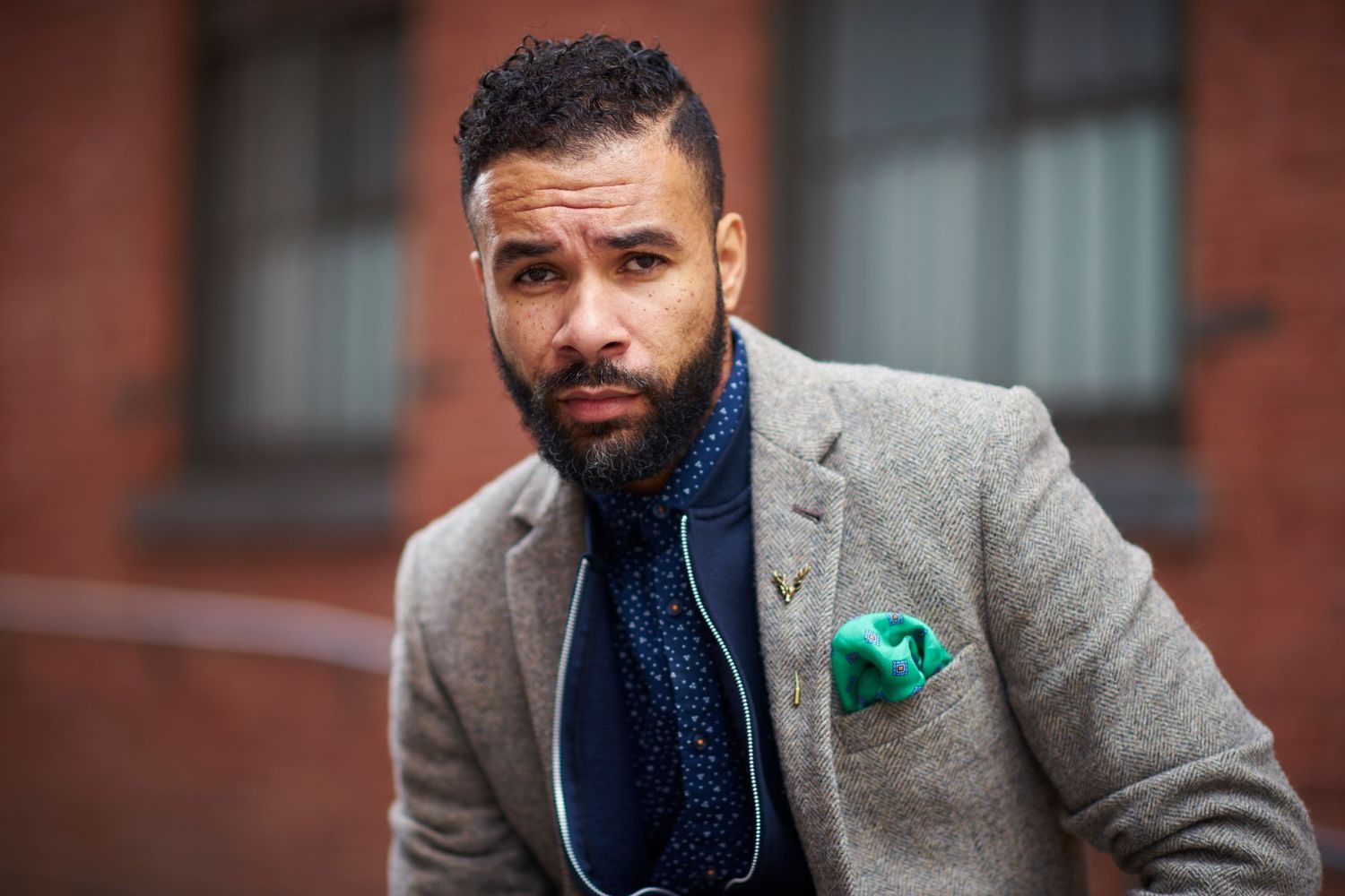 A well-dressed individual wearing a gray blazer with polka dot pocket square and navy patterned shirt poses outdoors.