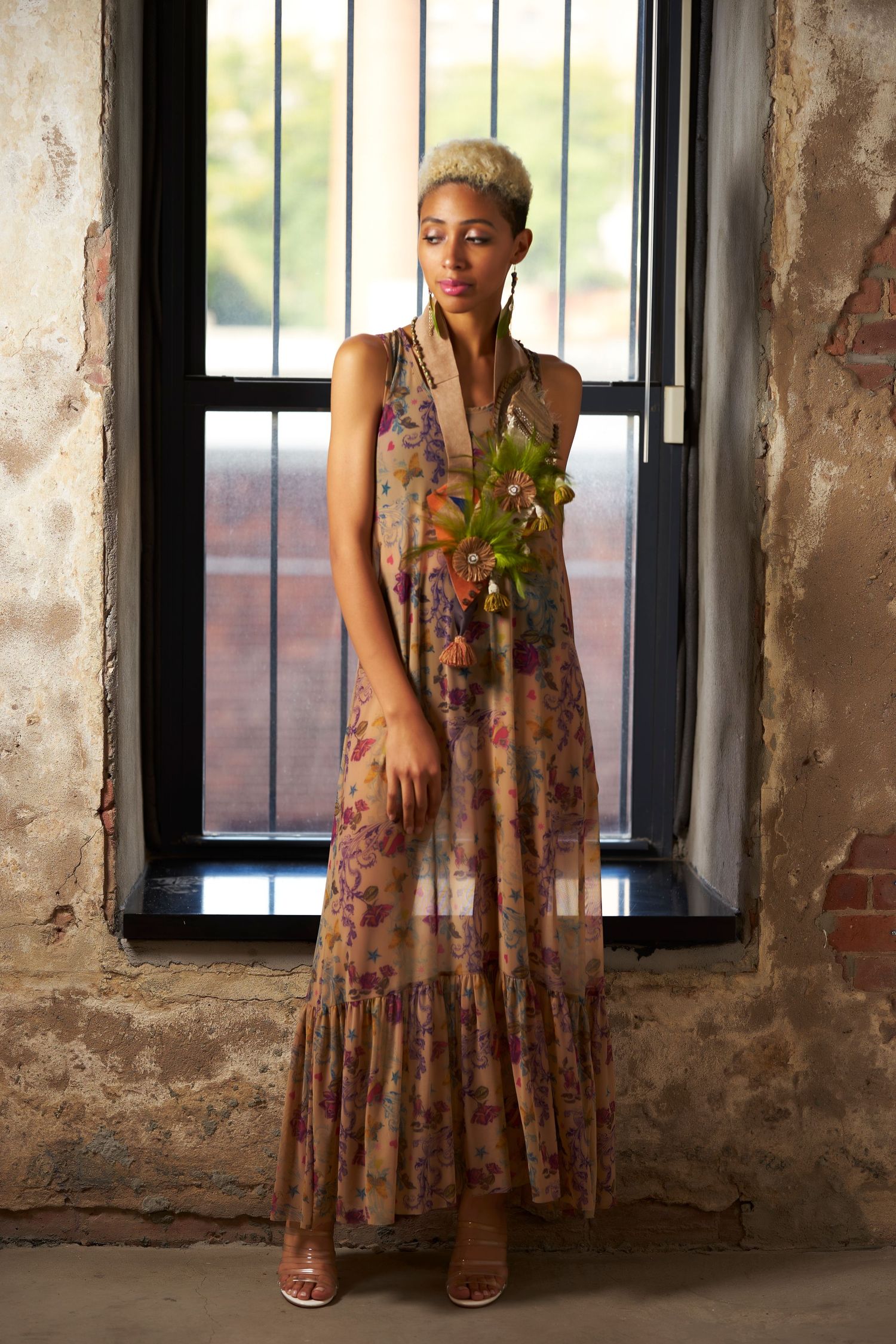 Woman in floral maxi dress poses in industrial space with exposed brick walls and black-framed windows.