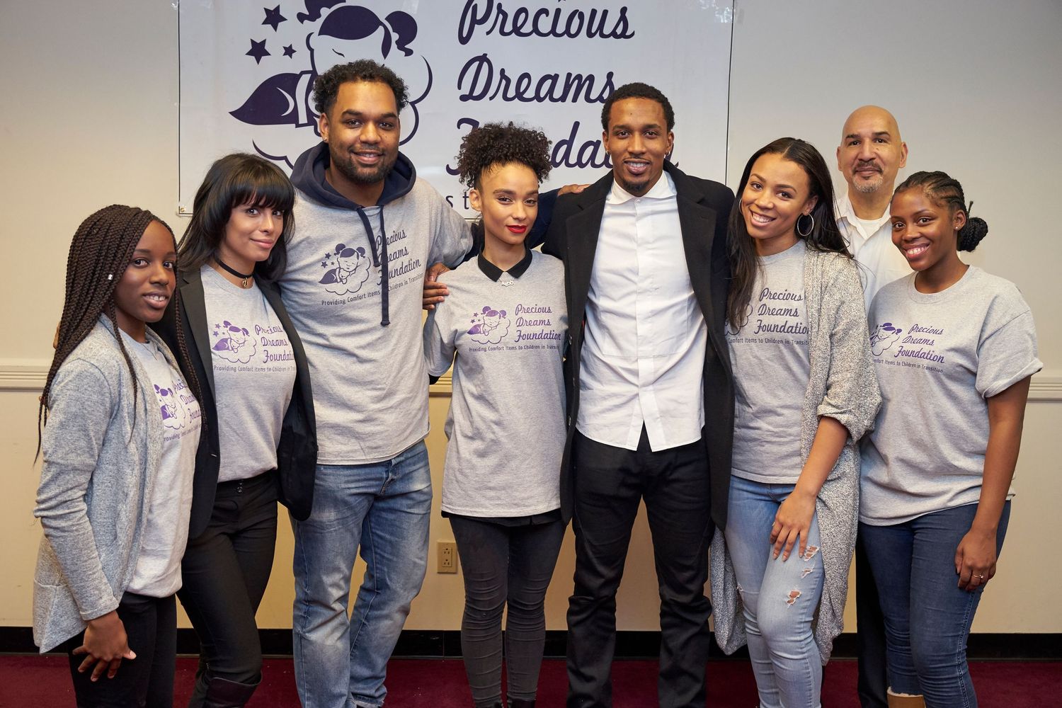 Group of people wearing matching gray sweatshirts and t-shirts pose together at an event with a Precious Dreams banner.