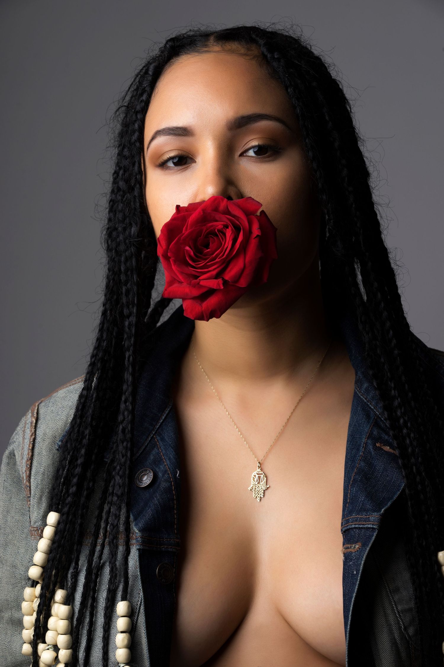 Artistic portrait featuring long braids, denim jacket and vibrant red rose against gray background.